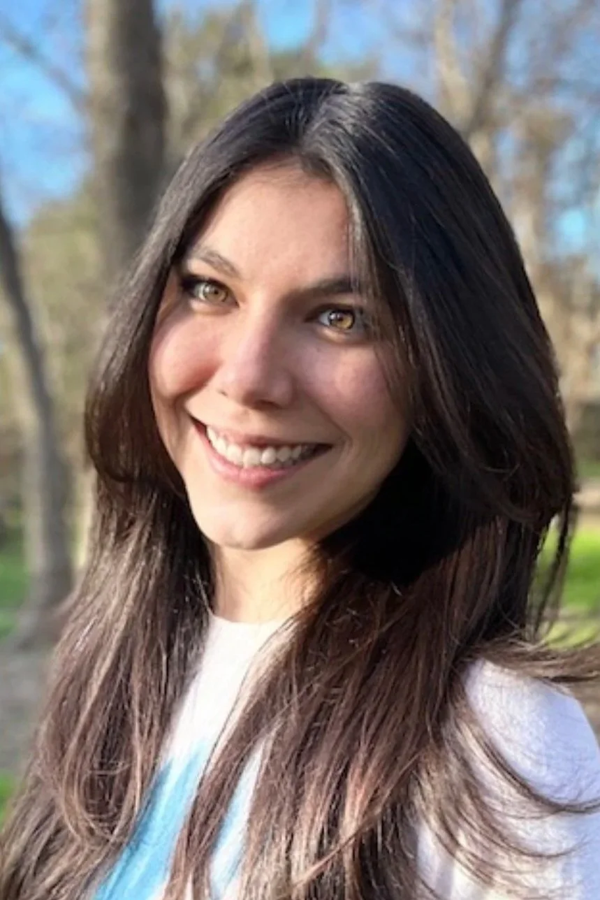 A woman with long dark hair and light-colored eyes smiling outdoors in a wooded area during daytime.