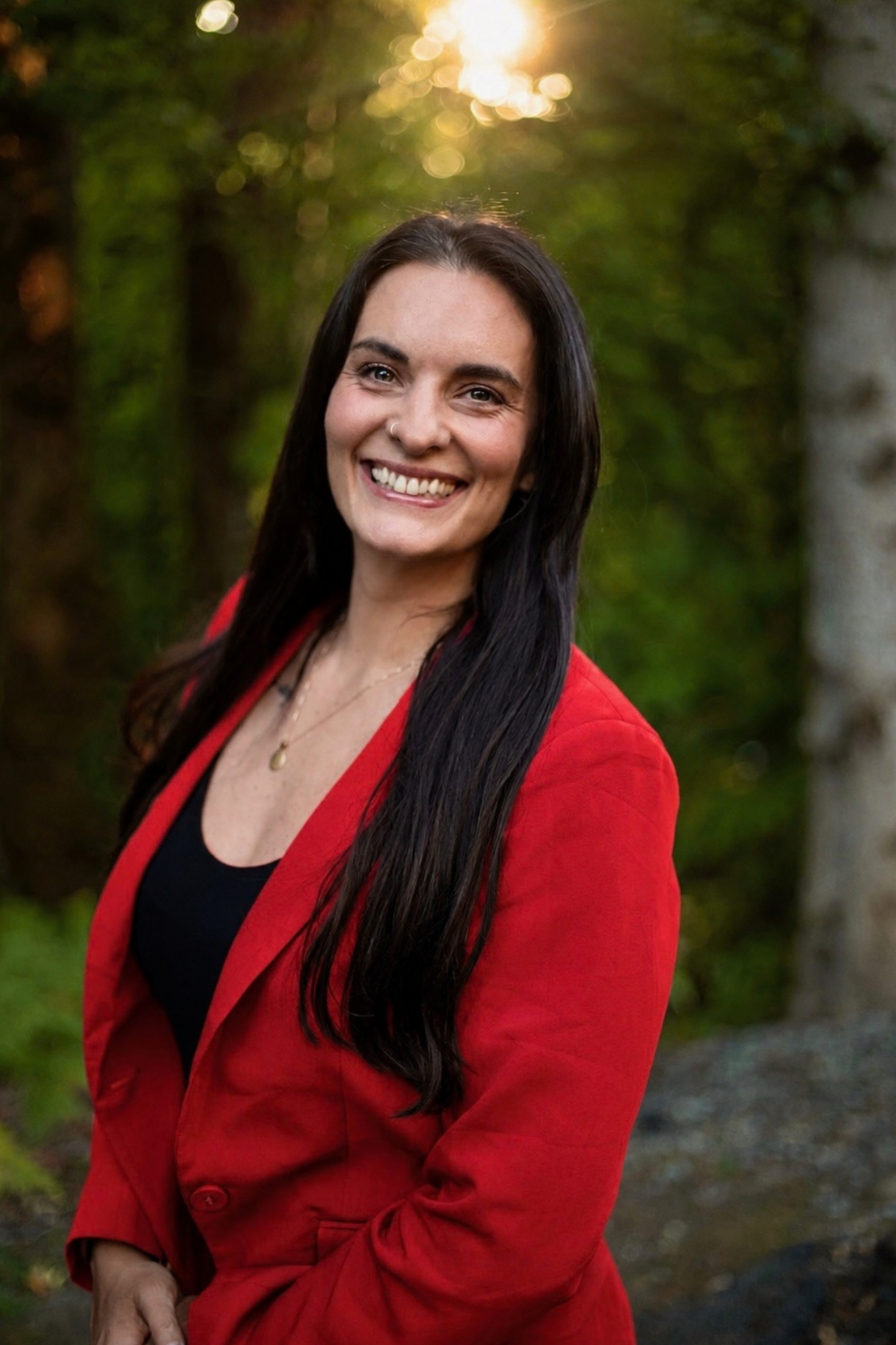 A woman with long dark hair, smiling, wearing a red jacket over a black top, standing outdoors with a blurred green and brown natural background, and sunlight visible behind her.