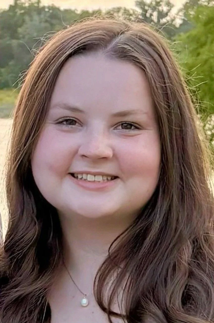A young woman with long brown hair and fair skin smiling outdoors.