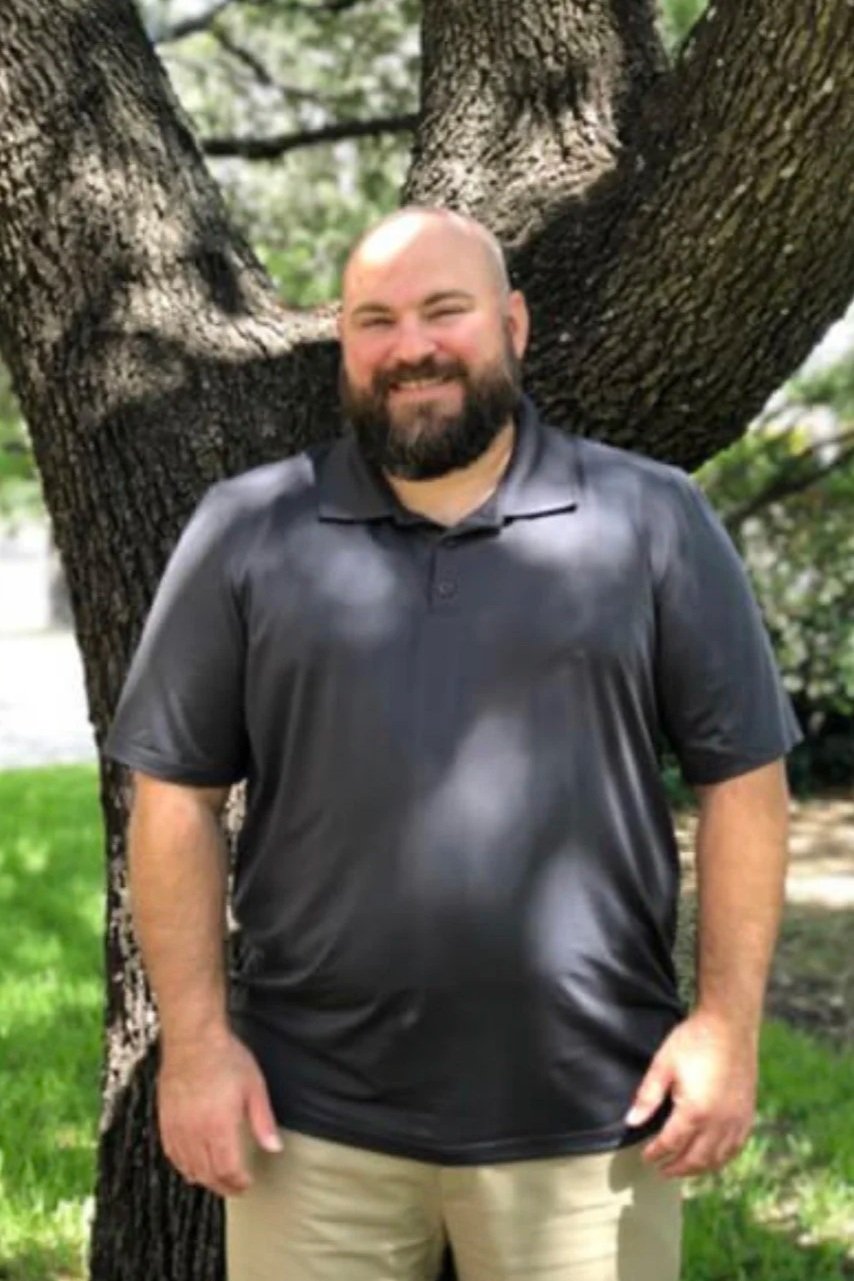 A man with a beard wearing a black polo shirt and beige pants standing outdoors in front of a large tree with textured bark, smiling.