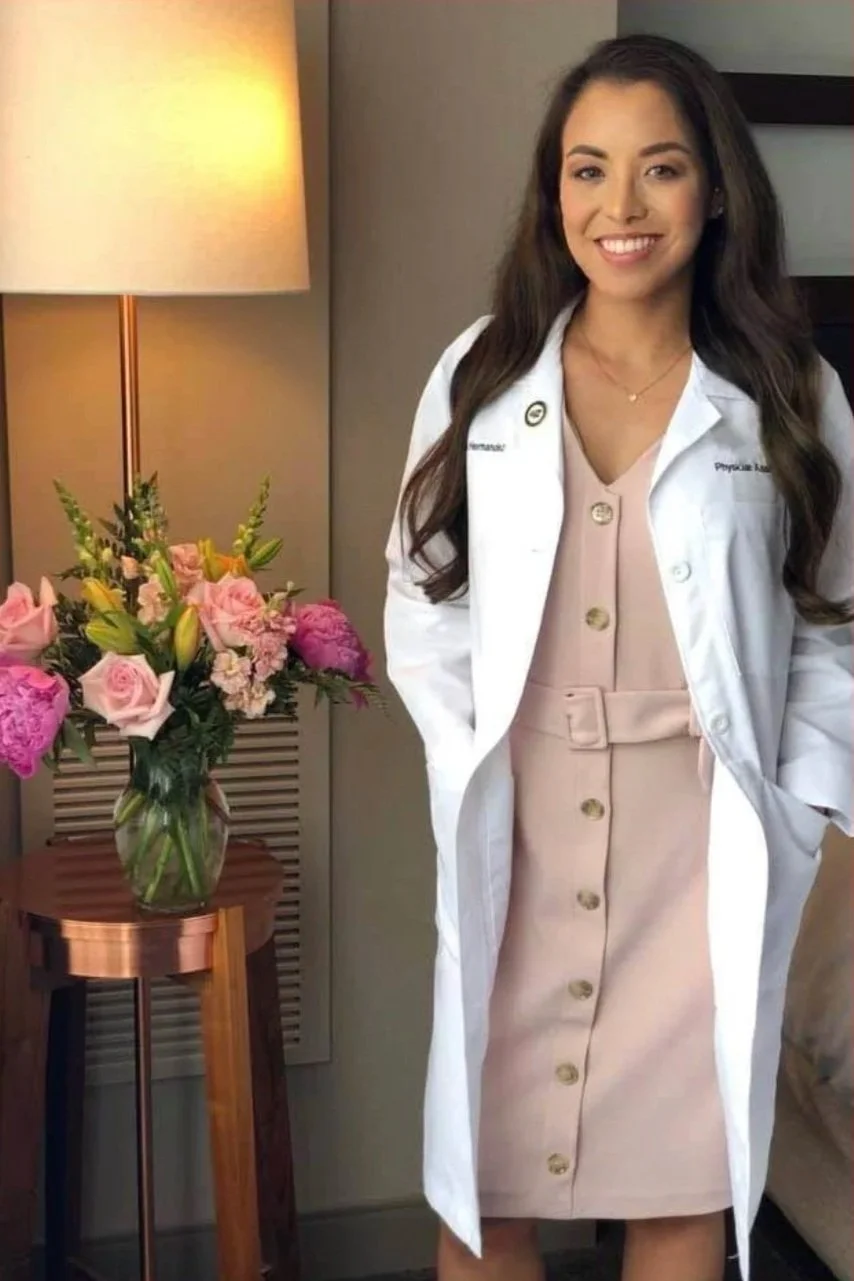 A woman with long brown hair wearing a white lab coat, standing next to a table with a vase of pink and peach flowers, smiling at the camera in a well-lit room.