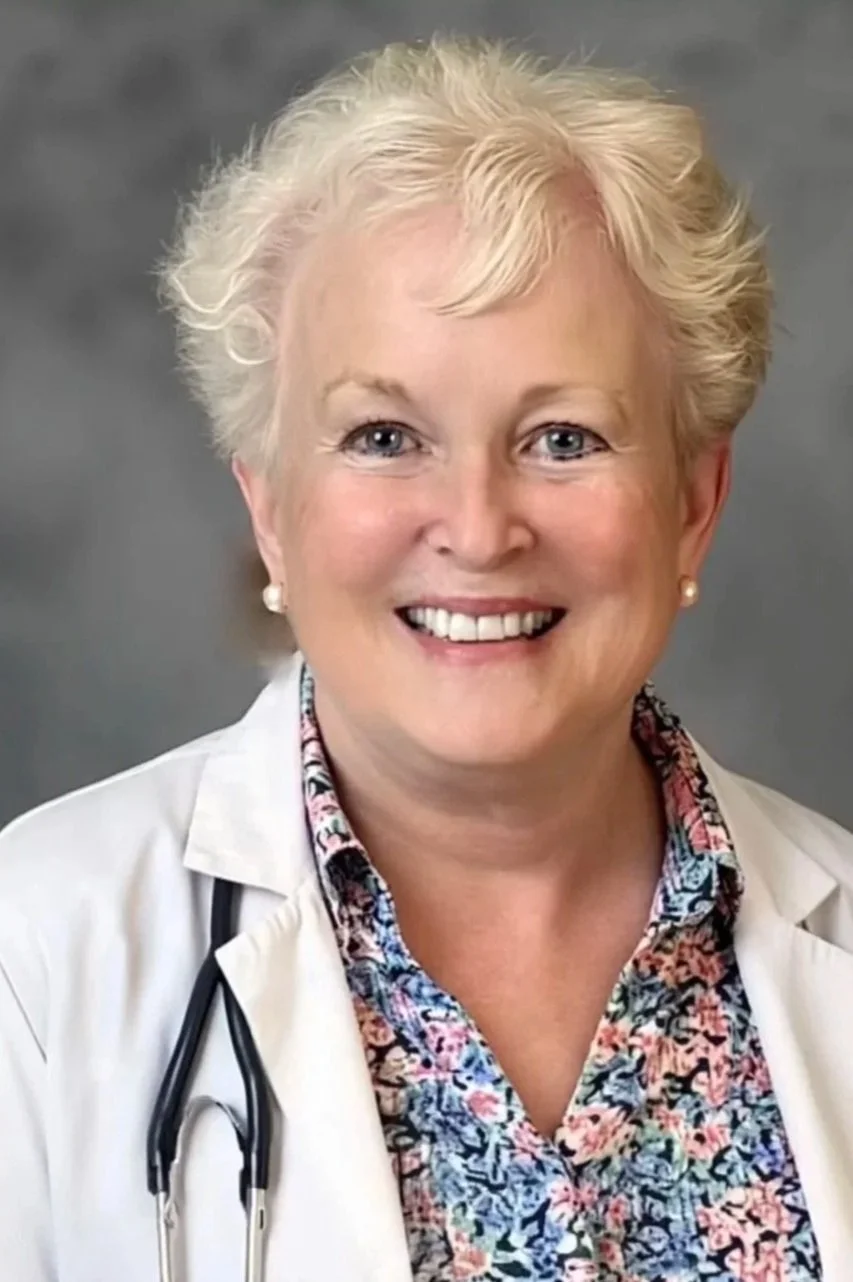 A middle-aged woman with short, curly blonde hair, wearing a white medical coat, a floral shirt, and pearl earrings, smiling in front of a gray background, with a stethoscope around her neck.