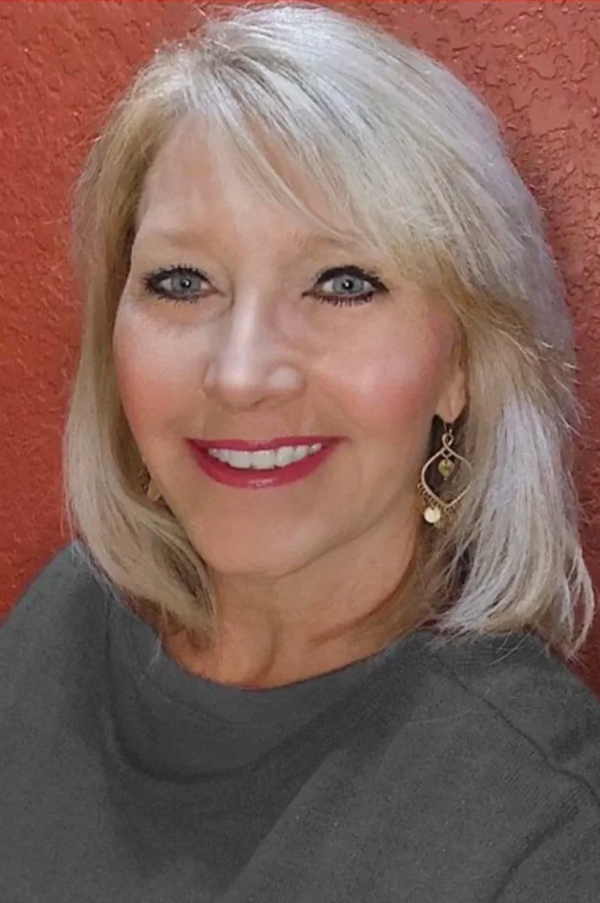 A middle-aged woman with shoulder-length blonde hair, blue eyes, and red lipstick, smiling and wearing a dark grey top and gold earrings, standing against a red textured wall.
