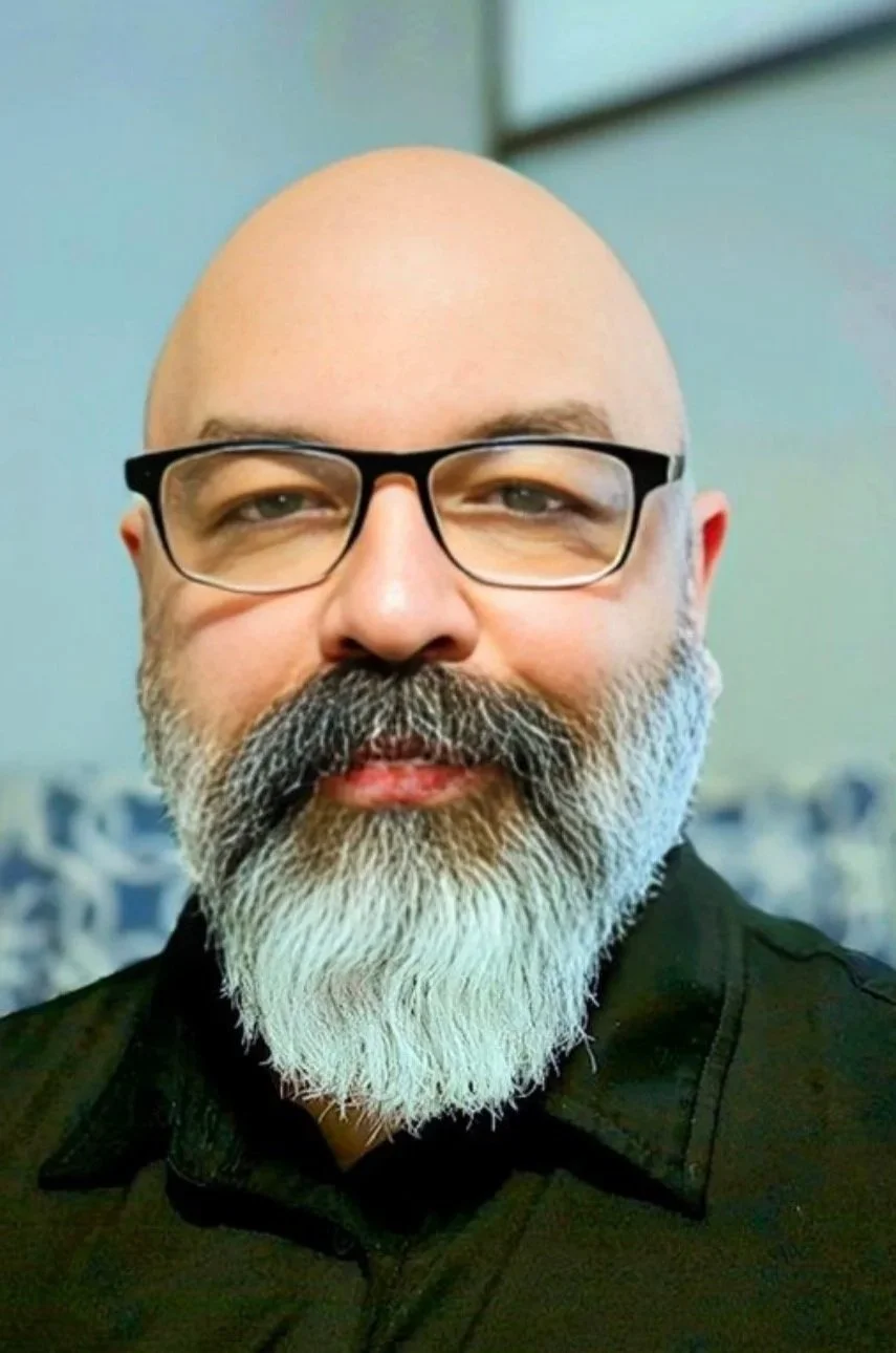 Close-up of a bald man with glasses, a full white beard, and a black shirt. He has a neutral expression and is indoors with a blurred background.