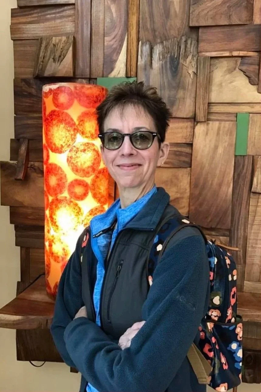 A woman wearing glasses, a blue jacket, and a backpack standing in front of a wooden wall and a glowing orange lamp.