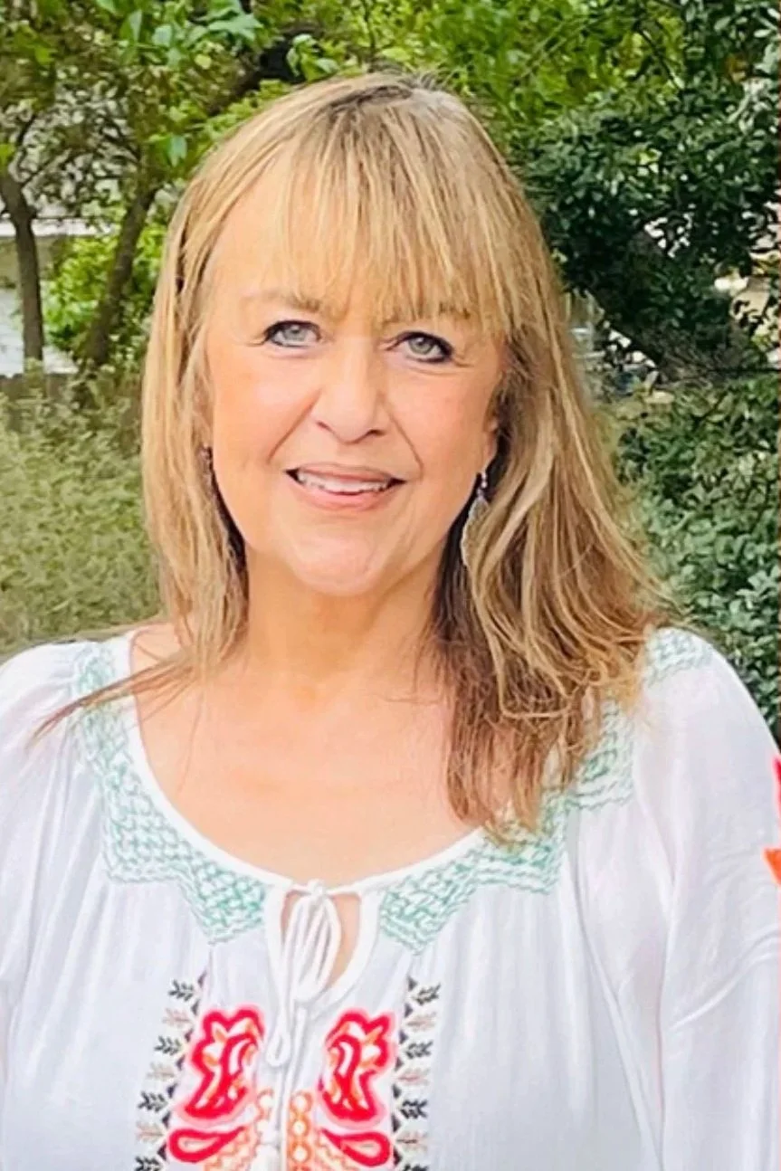 A woman with blonde hair smiling outdoors in a garden, wearing a white embroidered top.