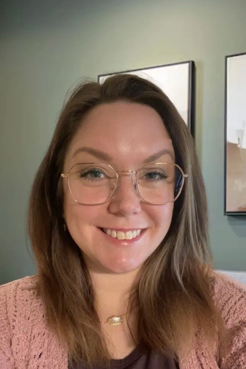 A woman with long brown hair and glasses smiling at the camera, indoors with framed pictures on the wall behind her.