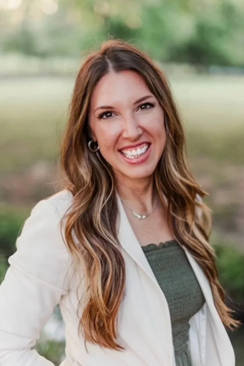 A woman with long wavy brown hair smiling outdoors.