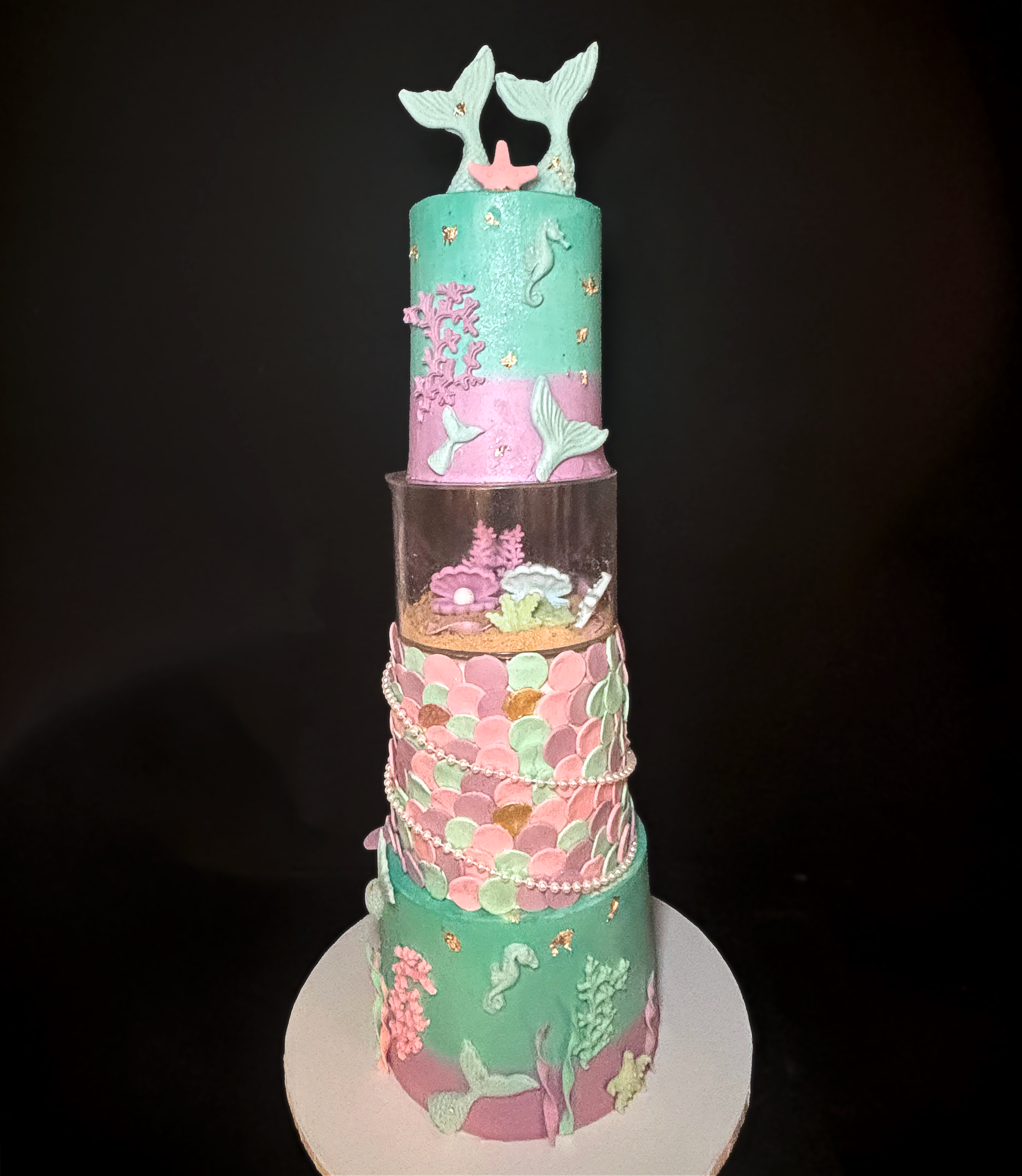 A tall, multi-tiered cake decorated with mermaid and underwater theme. The cake features pastel colors including pink, green, and blue, with decorative sea creatures, coral, and shells. The top has mermaid tails, with mermaid figures and fish around 