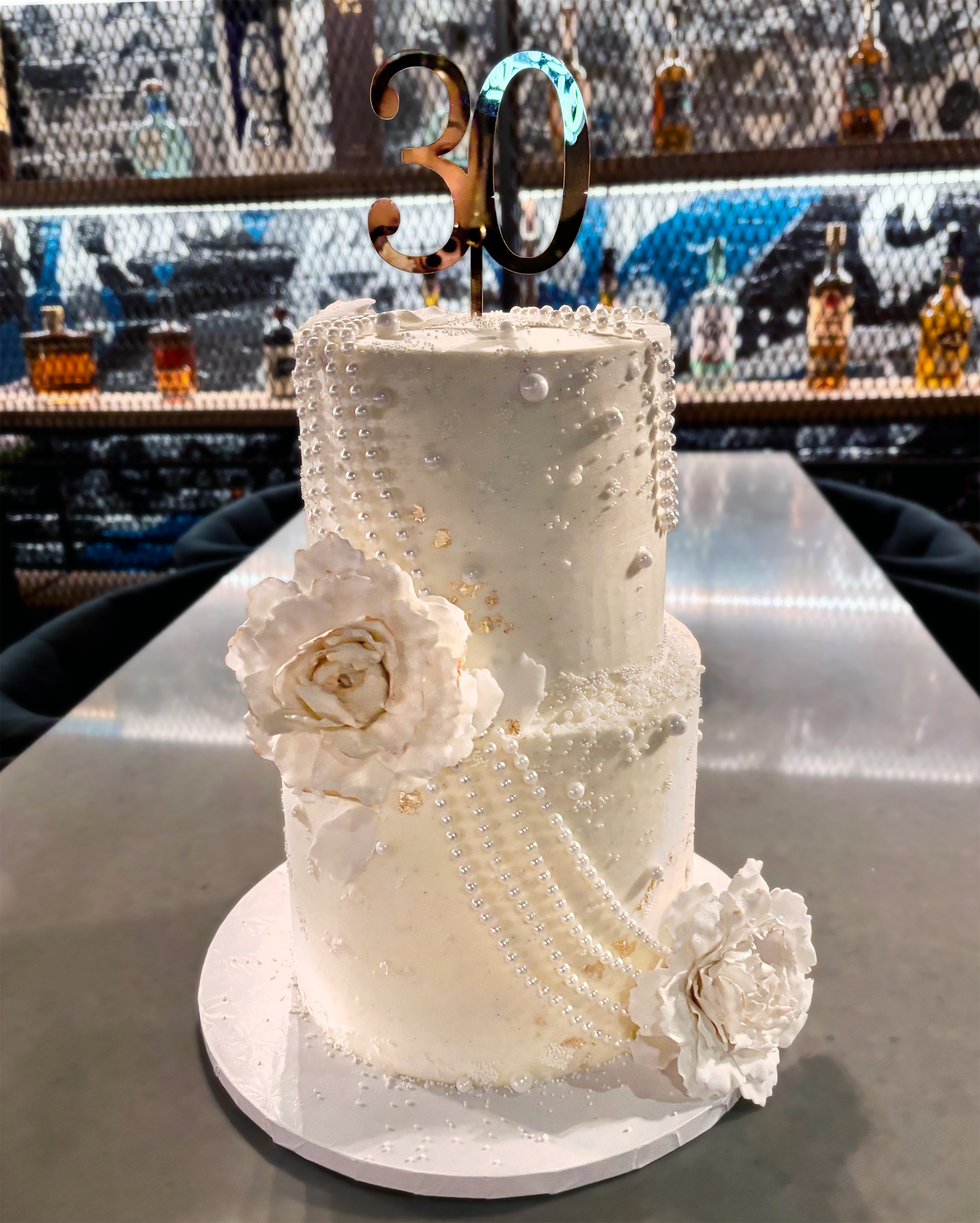 A white birthday cake decorated with pearls, a white rose, and a large gold number 30 cake topper, on a white cake board, with a bar background in the distance.