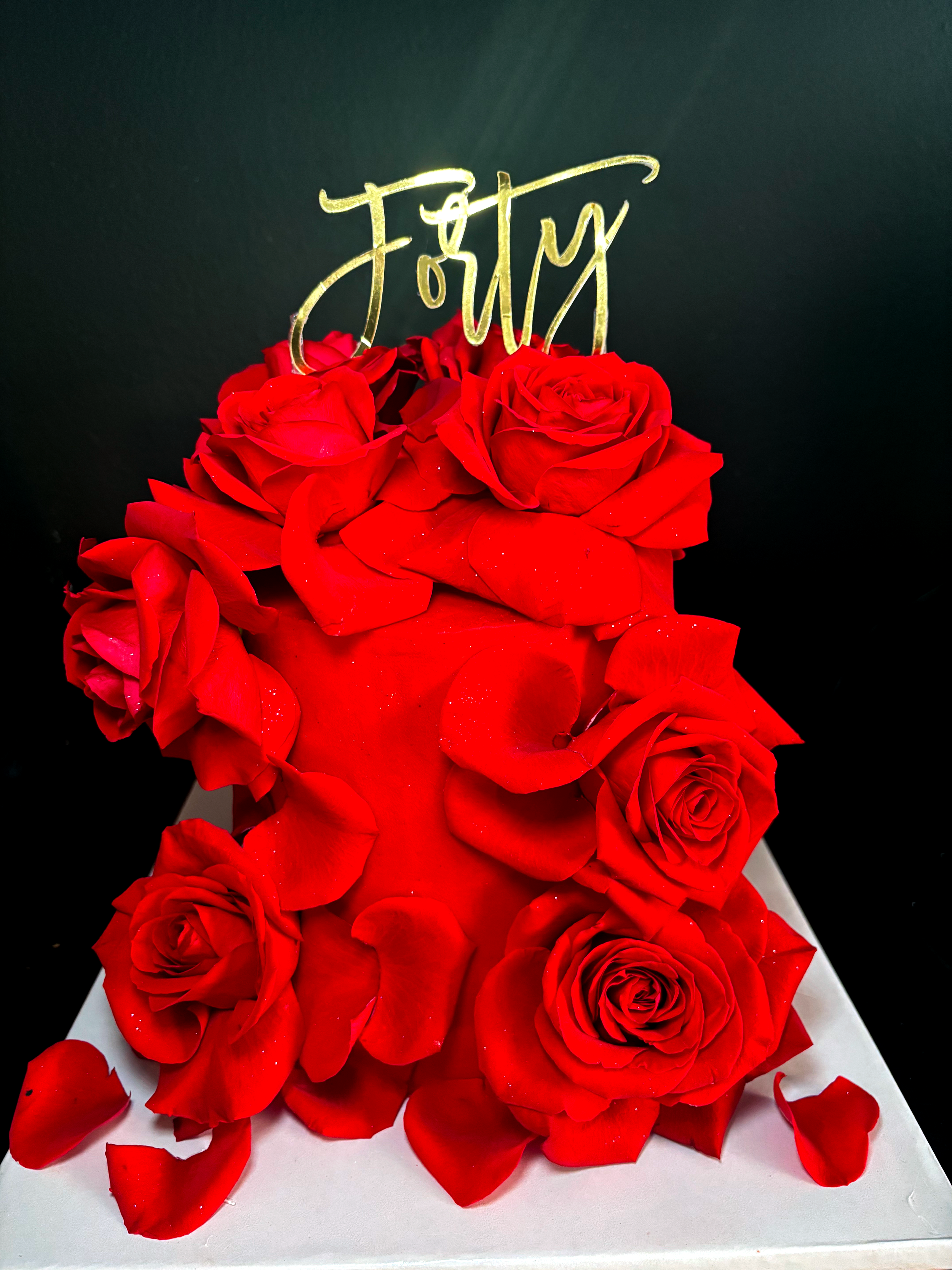 Red edible rose-covered cake with a "Forty" topper on a white surface against a dark background.