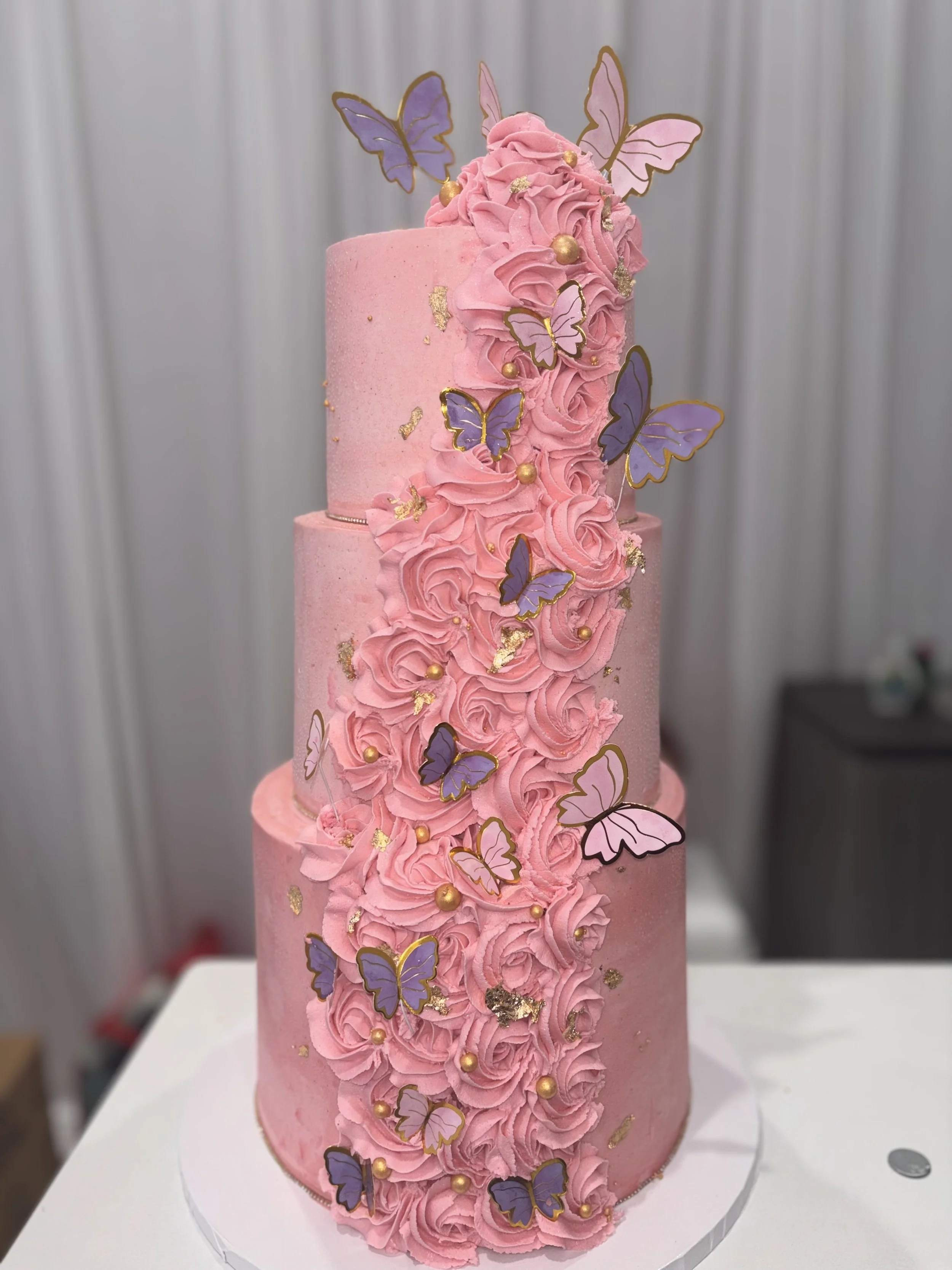 Three-tier pink cake decorated with buttercream roses, purple butterfly toppers, and gold accents.