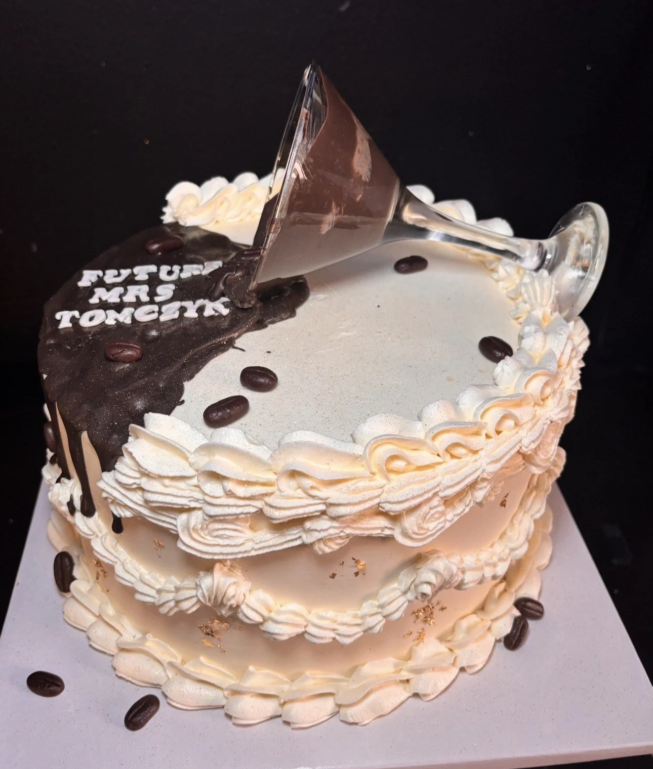 A birthday cake with white and chocolate frosting, decorated with coffee beans, gold flakes, and a message that reads 'Future MHS Today' in white icing. A glass upside-down martini glass and a piece of chocolate are on top of the cake.