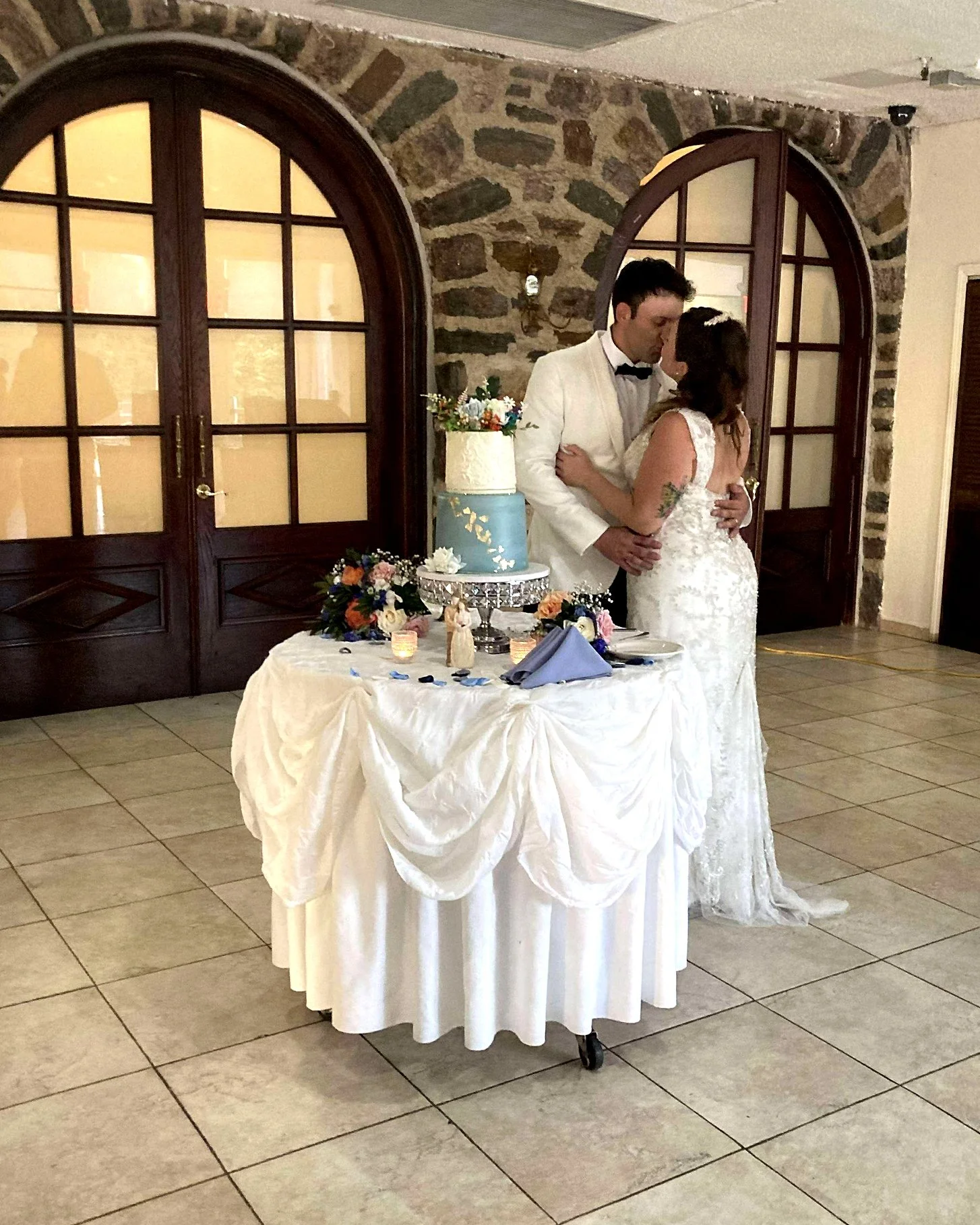 A wedding reception with a bride and groom embracing behind a decorated cake table in a rustic venue with stone walls and wooden arch windows.