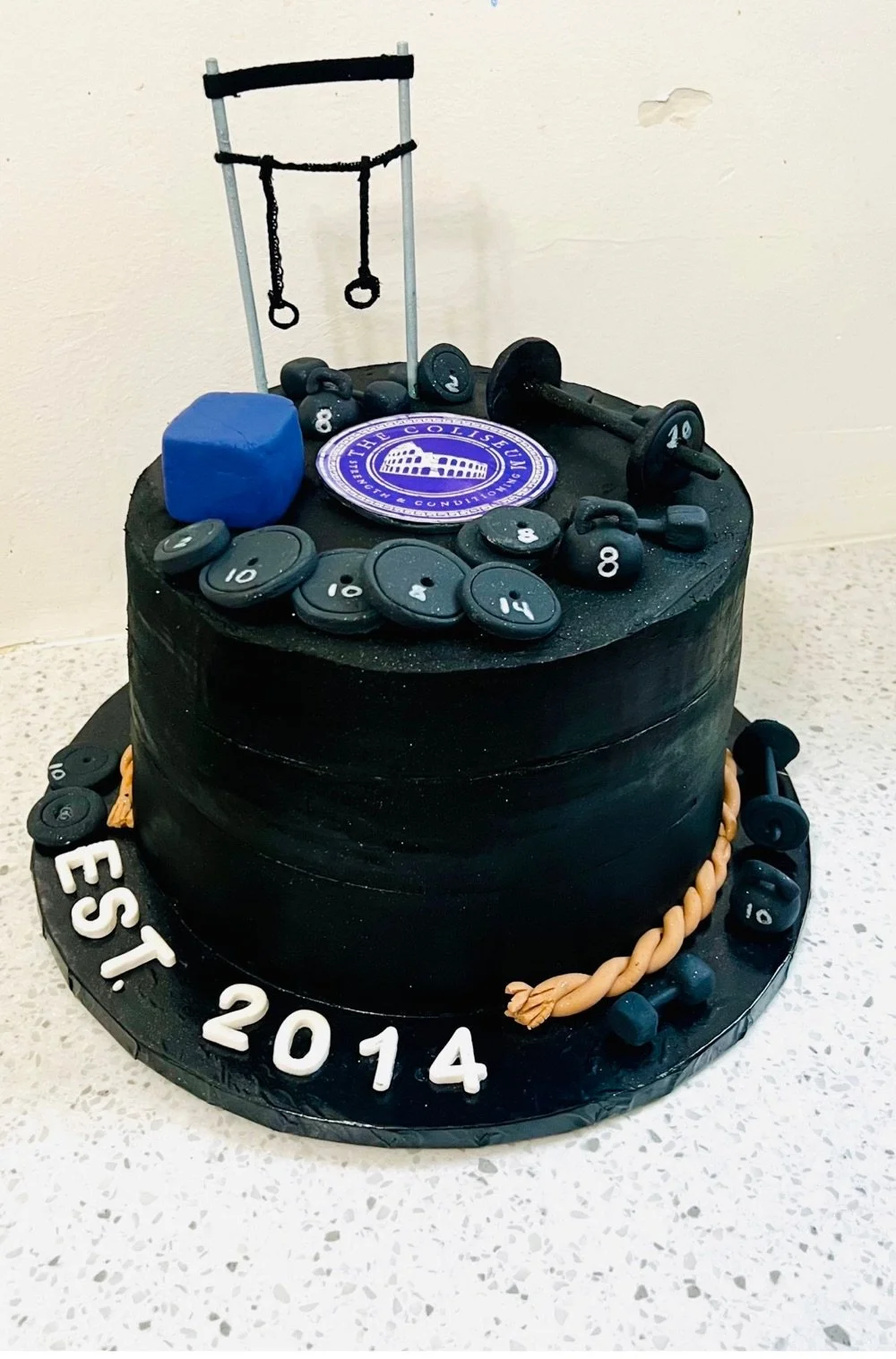 A black birthday cake decorated with fitness-themed edible decorations, including miniature dumbbells, weights, gym equipment, and a logo. The cake has white and orange cord-like embellishments, and the text "EST. 2014" on the base.