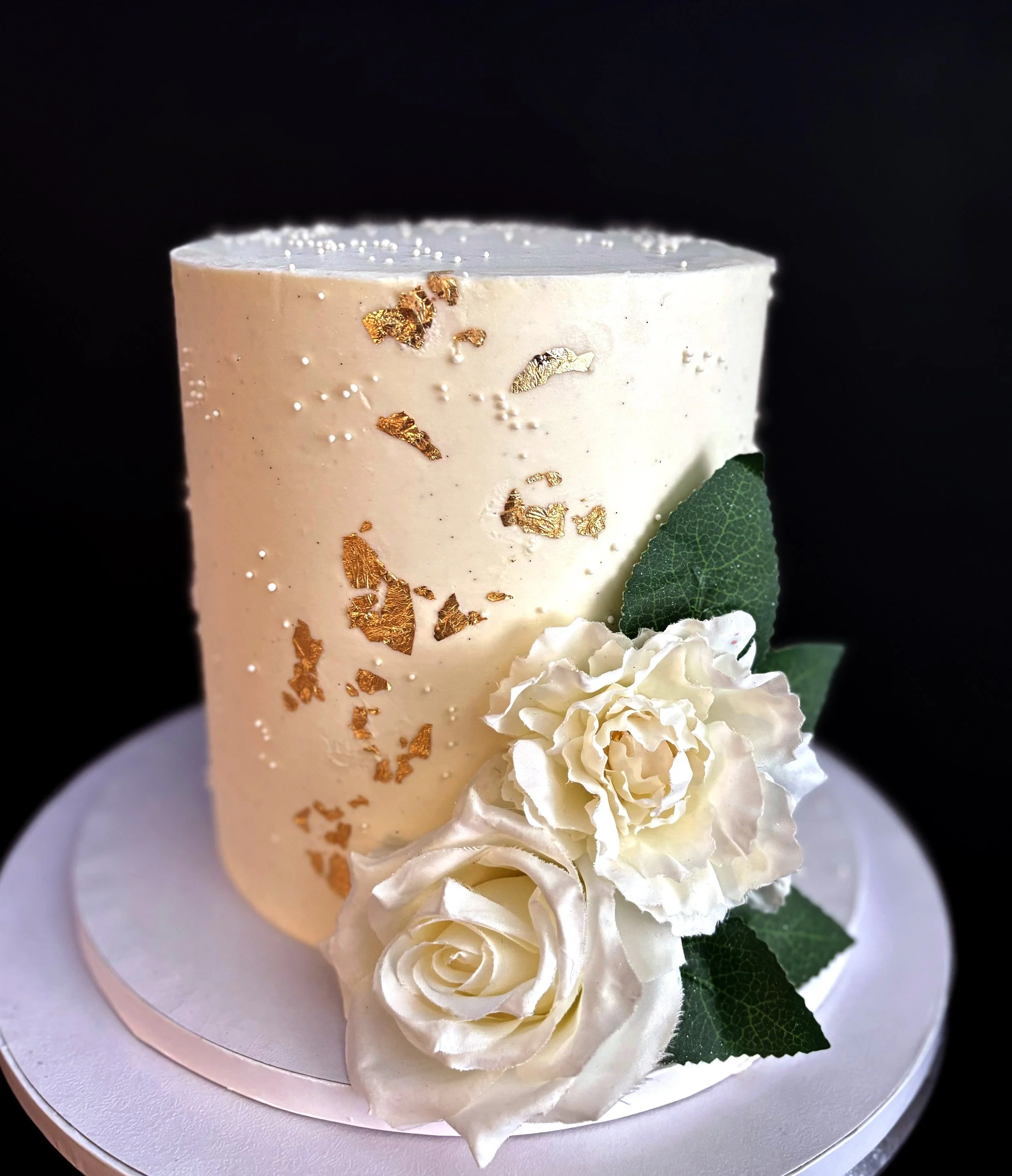 Elegant white cake decorated with gold leaf and tiny white sugar pearls, adorned with white roses and green leaves, against a black background.