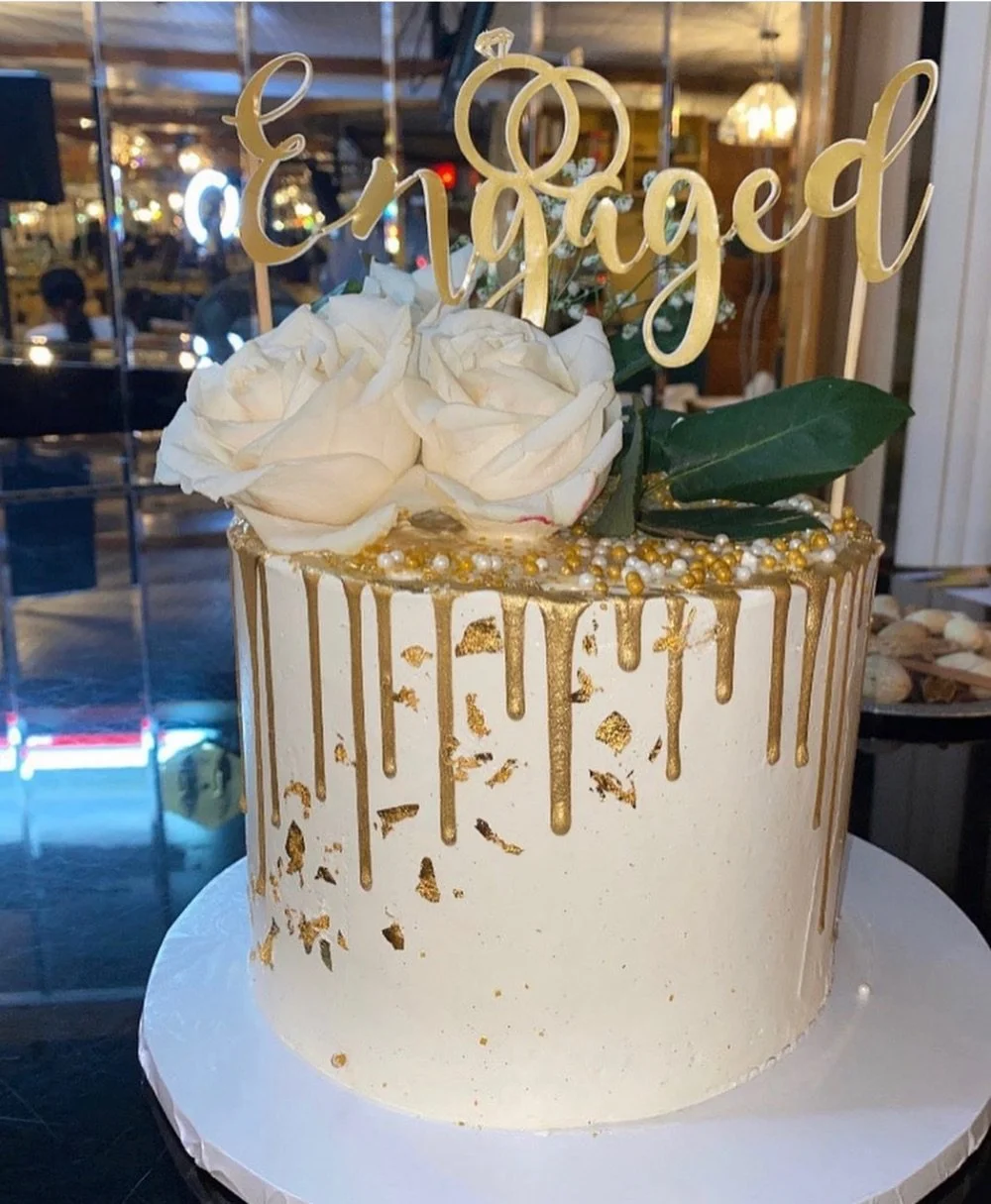 A white cake with gold drip decoration, white roses, and a gold "Engaged" topper for an engagement celebration.