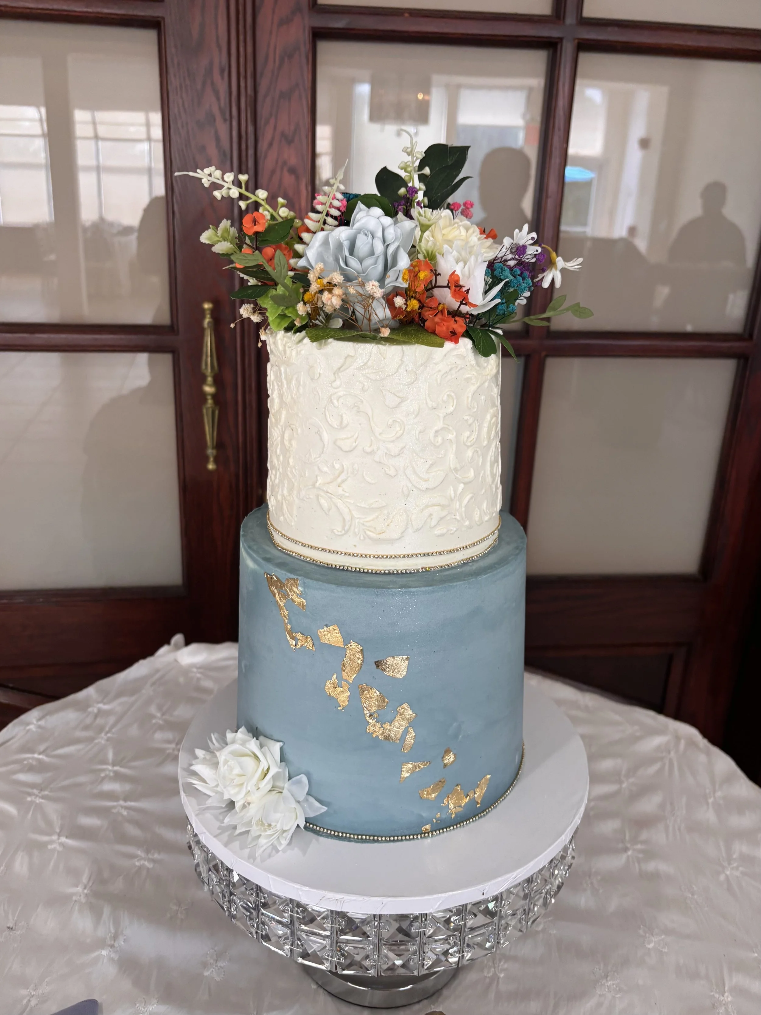 A three-tiered wedding cake with a white top tier decorated with textured icing and fresh flowers, a middle tier with a silver satin finish and gold accents, and a bottom tier with a blue satin finish and gold leaf designs, placed on a decorative sta