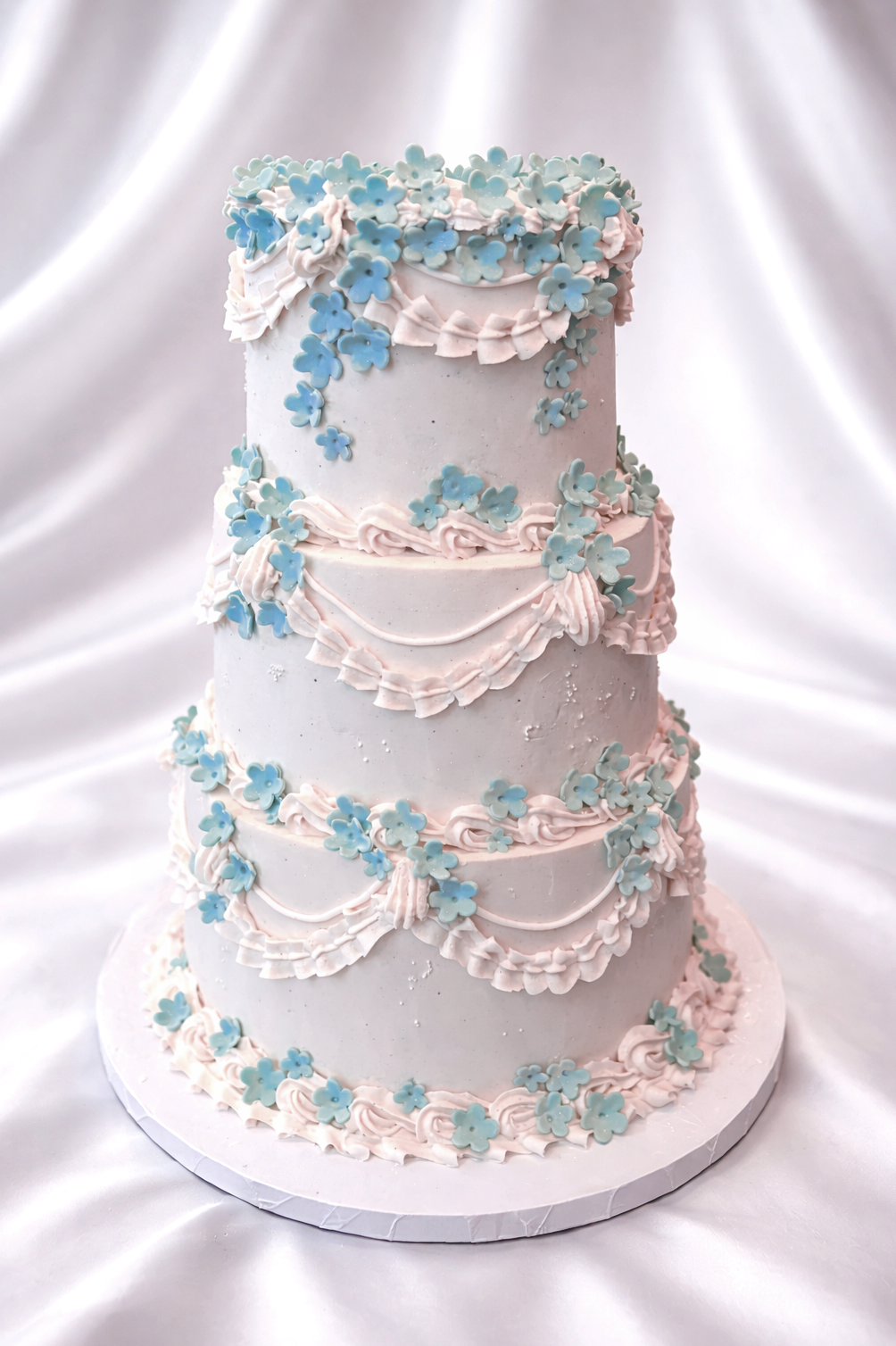 Three-tiered wedding cake decorated with blue flowers and pink icing on a white satin background.
