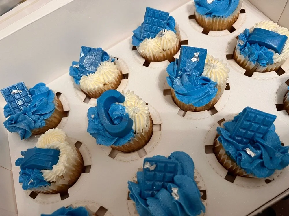 Blue and white decorated cupcakes with chocolate pieces on top, arranged in a white box.