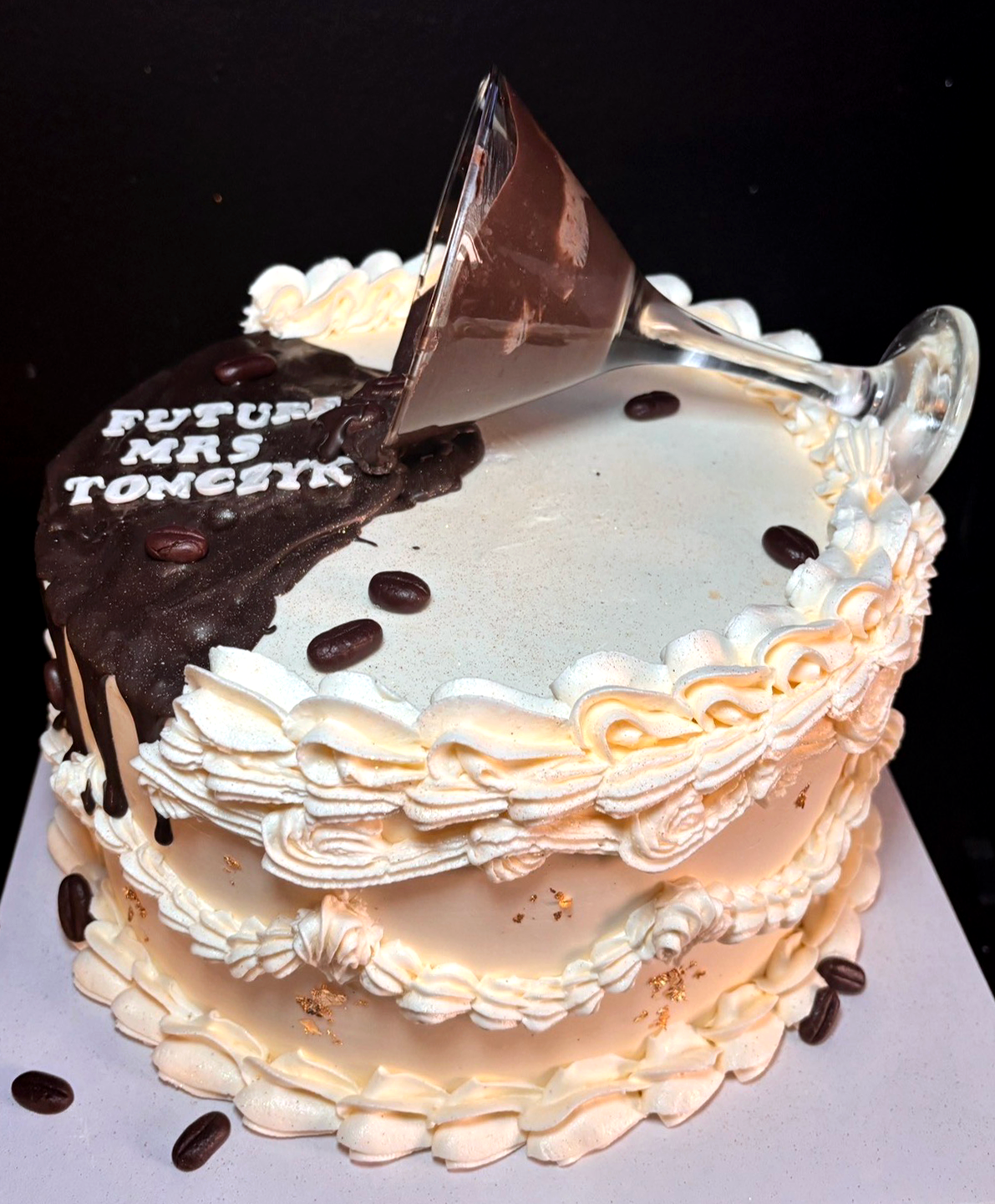 A birthday cake decorated with white frosting, topped with a large glass of wine on its side, and decorated with coffee beans and gold flakes, with the message 'FUTURE MRS TOMEK' written on a chocolate piece.