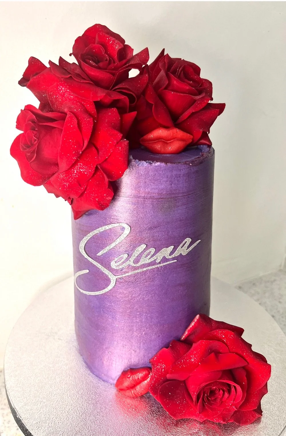 A purple cylindrical cake decorated with red roses and lips, with the name 'Selena' written on it, placed on a round silver cake board.