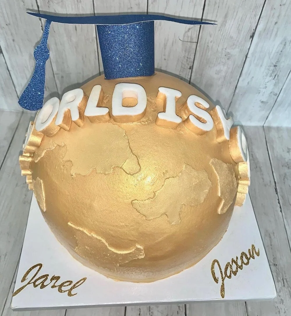 A globe-shaped cake with a gold finish, decorated with a blue graduation cap and the words 'WORLD ISY' on top; the cake is on a white base with gold cursive writing of the names Jarrel and Jaaran.