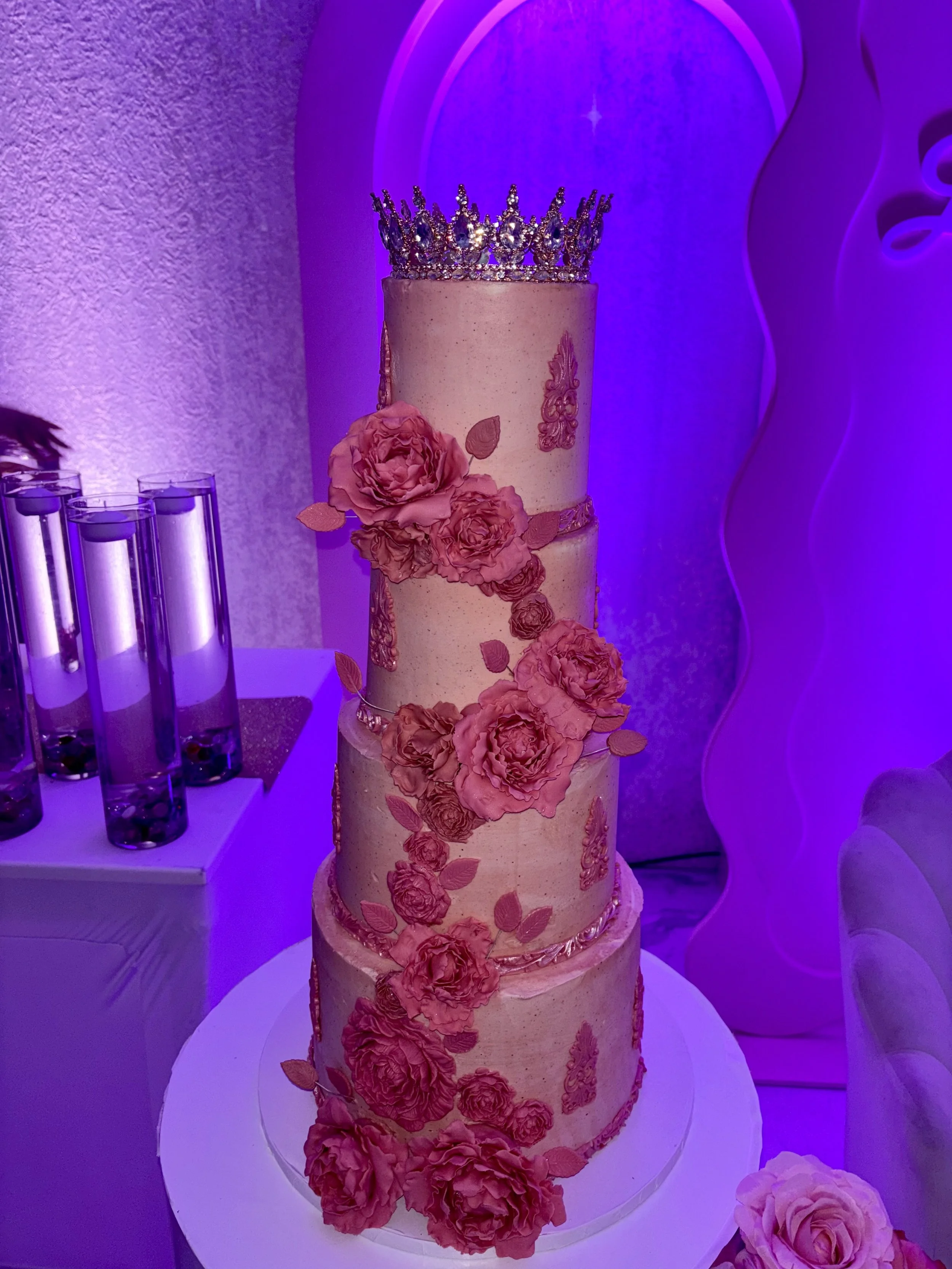 Three-tiered wedding cake with pink floral decorations and a silver tiara on top, placed on a white stand.