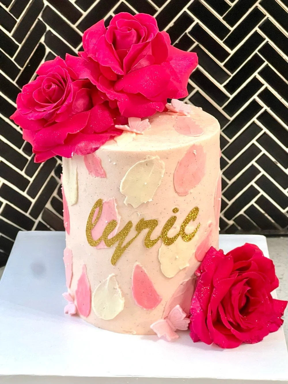 A pink birthday cake decorated with pink and cream petal-like decorations, pink roses on top and beside the cake, and the name 'Lynie' written in gold glitter. The background features a black and white herringbone pattern.