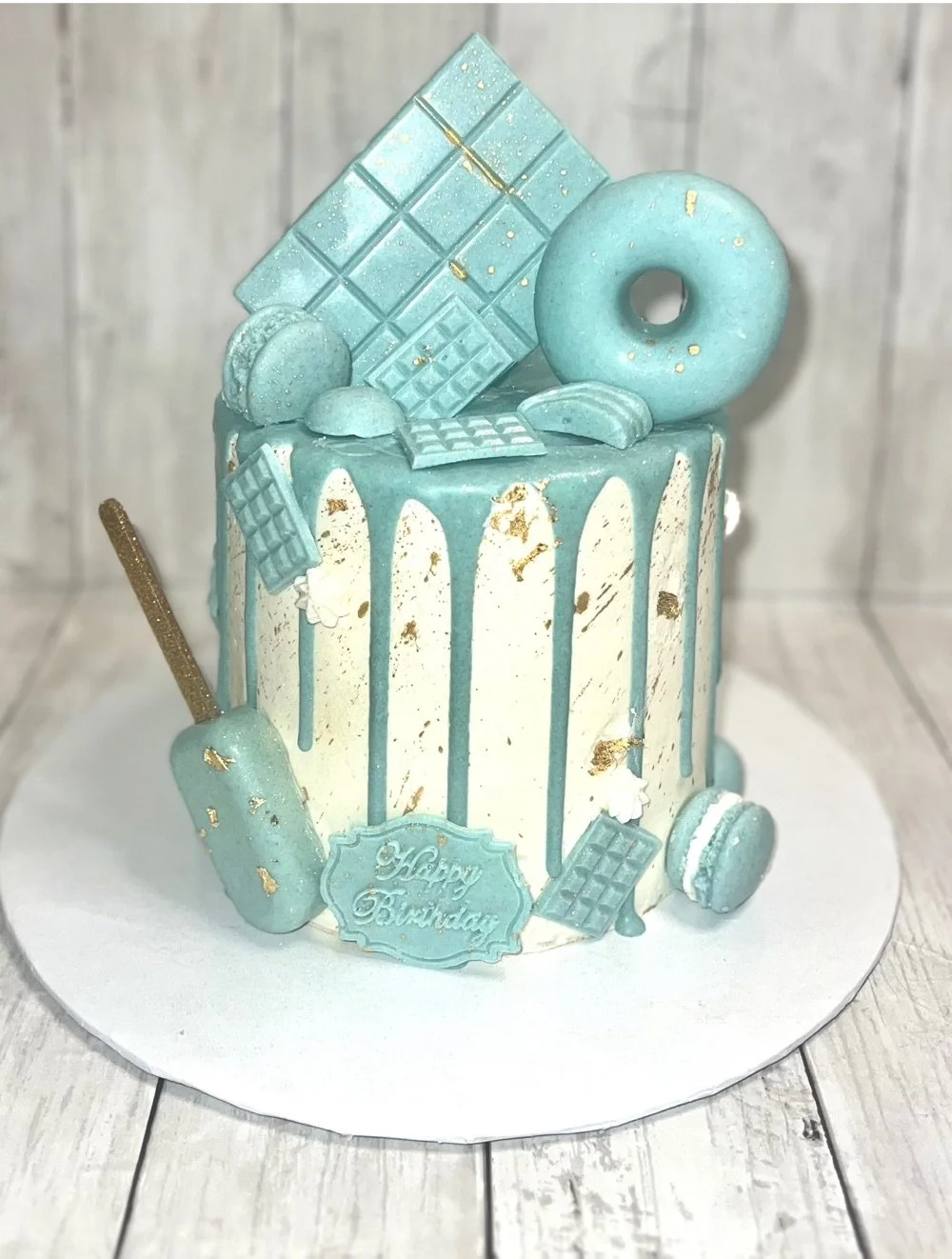 A tall birthday cake decorated with white and light blue icing, gold accents, and topped with blue chocolate pieces, macarons, and a donut. It features a "Happy Birthday" message on a blue plaque.