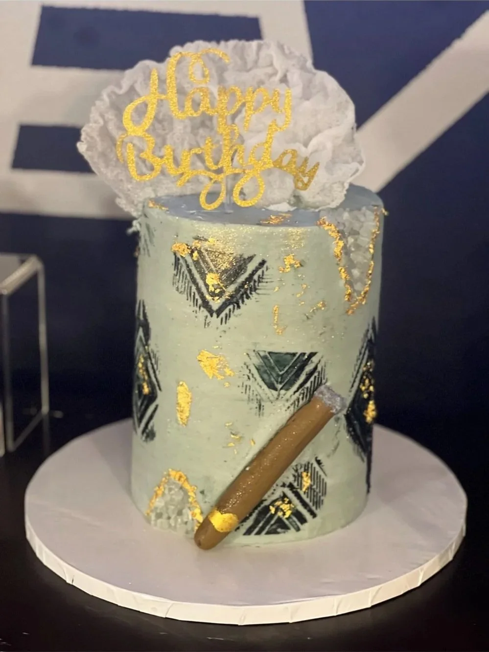 A birthday cake decorated with gold foil accents, black and white geometric patterns, a white textured topper with gold text reading 'Happy Birthday,' and a brown rolled wafer stick on the side.