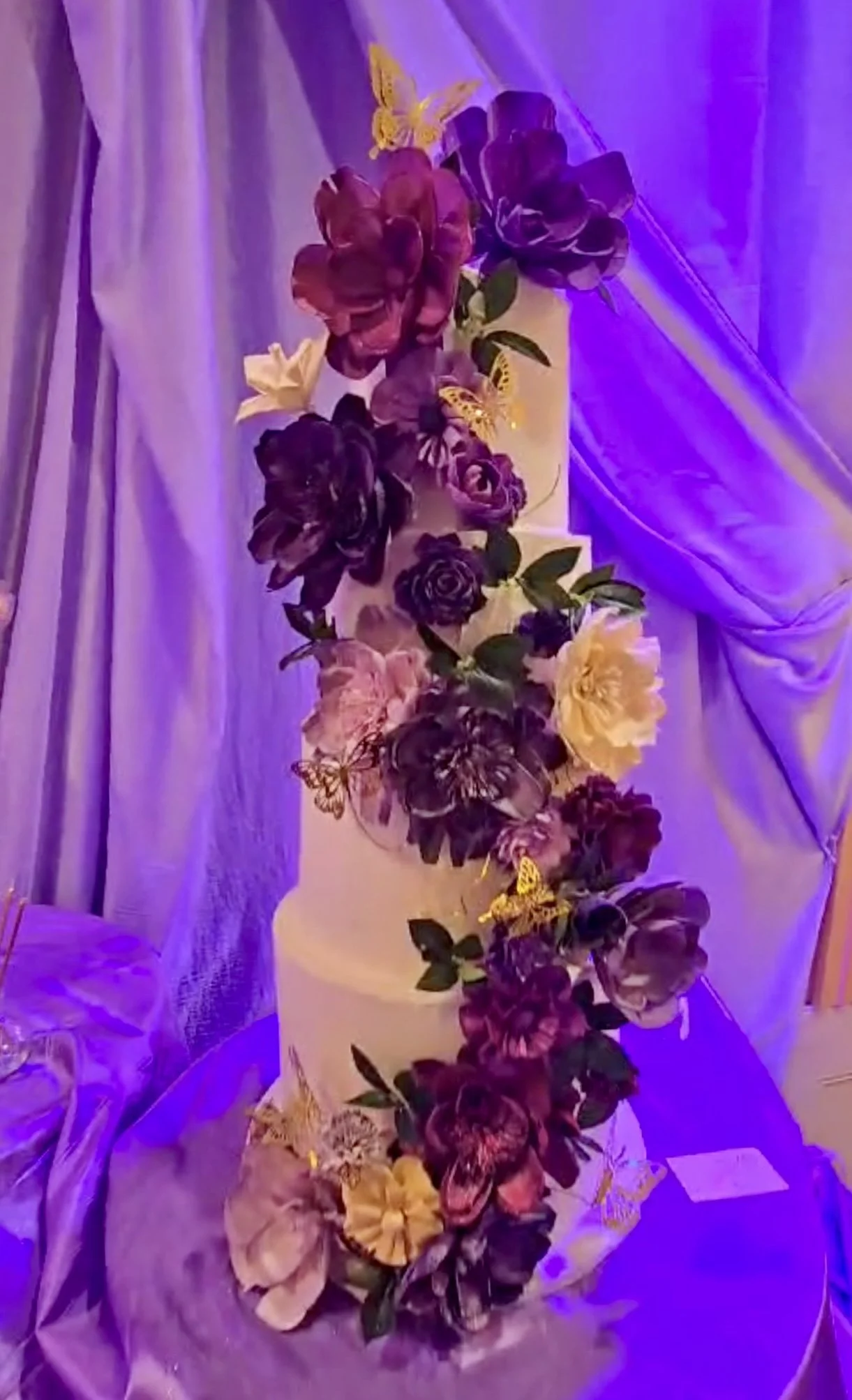 A tall white wedding cake decorated with purple, yellow, and blush flowers, including roses and orchids, along with yellow butterflies, set on a table with purple lighting and drapes in the background.