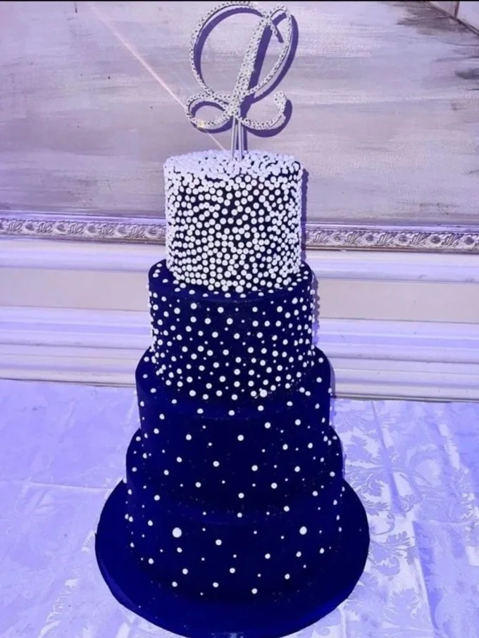 A four-tier black and white birthday cake with white polka dots, topped with a white cake topper in the shape of a stylized letter 'L' and a sword.