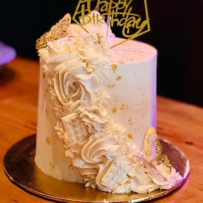 A birthday cake with white frosting, decorative cream swirls, gold accents, and a "Happy Birthday" topper, on a wooden surface.