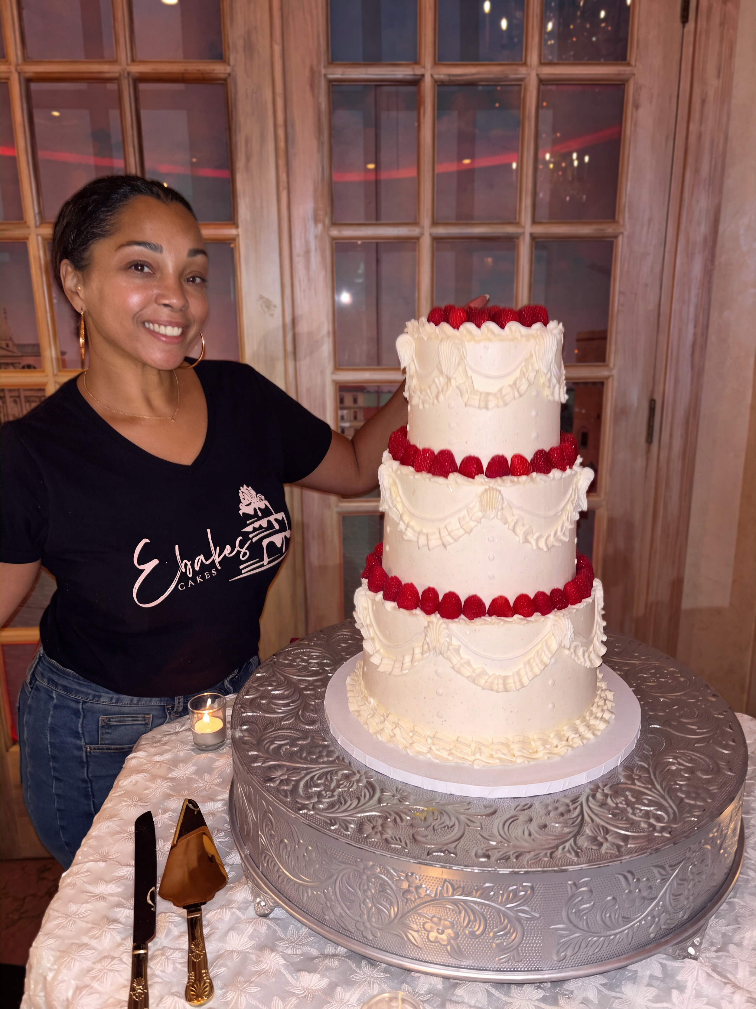 Woman smiling next to a tall, multi-tiered white cake decorated with strawberries and white frosting on a silver cake stand, with a wooden window in the background.