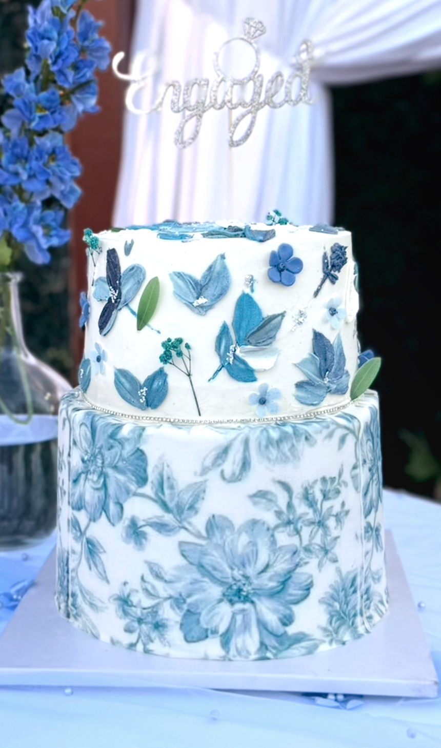 A two-tiered wedding cake with blue and white floral decorations and a silver 'engaged' cake topper. The cake is decorated with blue and white flowers and leaves, and is set on a table with a white tablecloth.