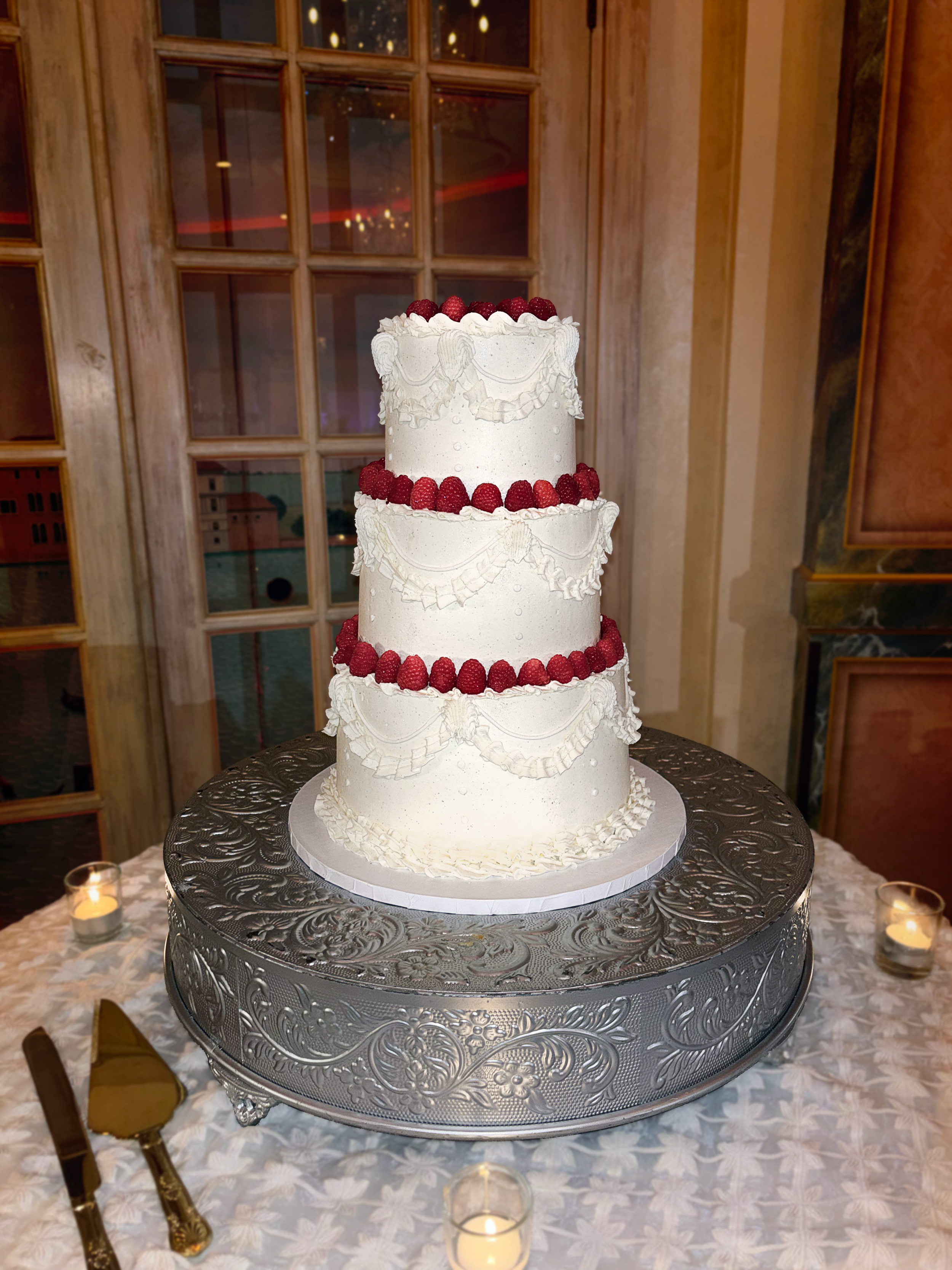 A large, multi-tiered white cake decorated with red raspberries and whipped cream, placed on a decorative silver cake stand in an elegant setting with candles and a patterned tablecloth.