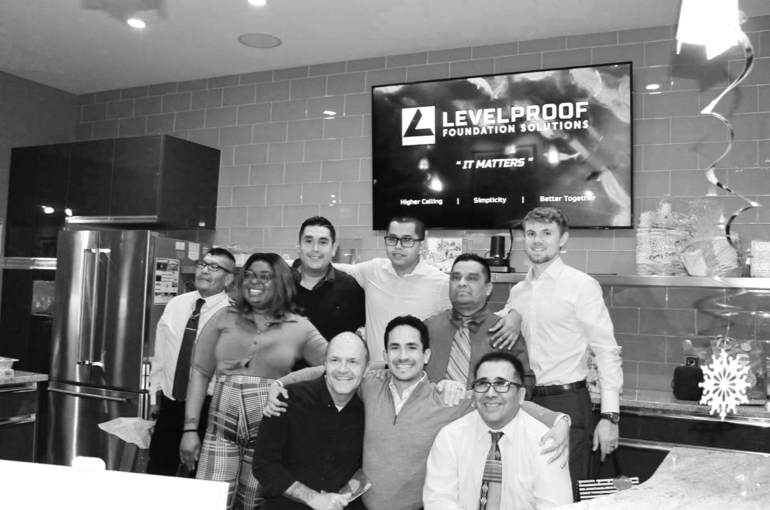 Group of ten diverse people posing for a photo in an office kitchen with a large screen displaying 'LevelProof Foundation Solutions'.