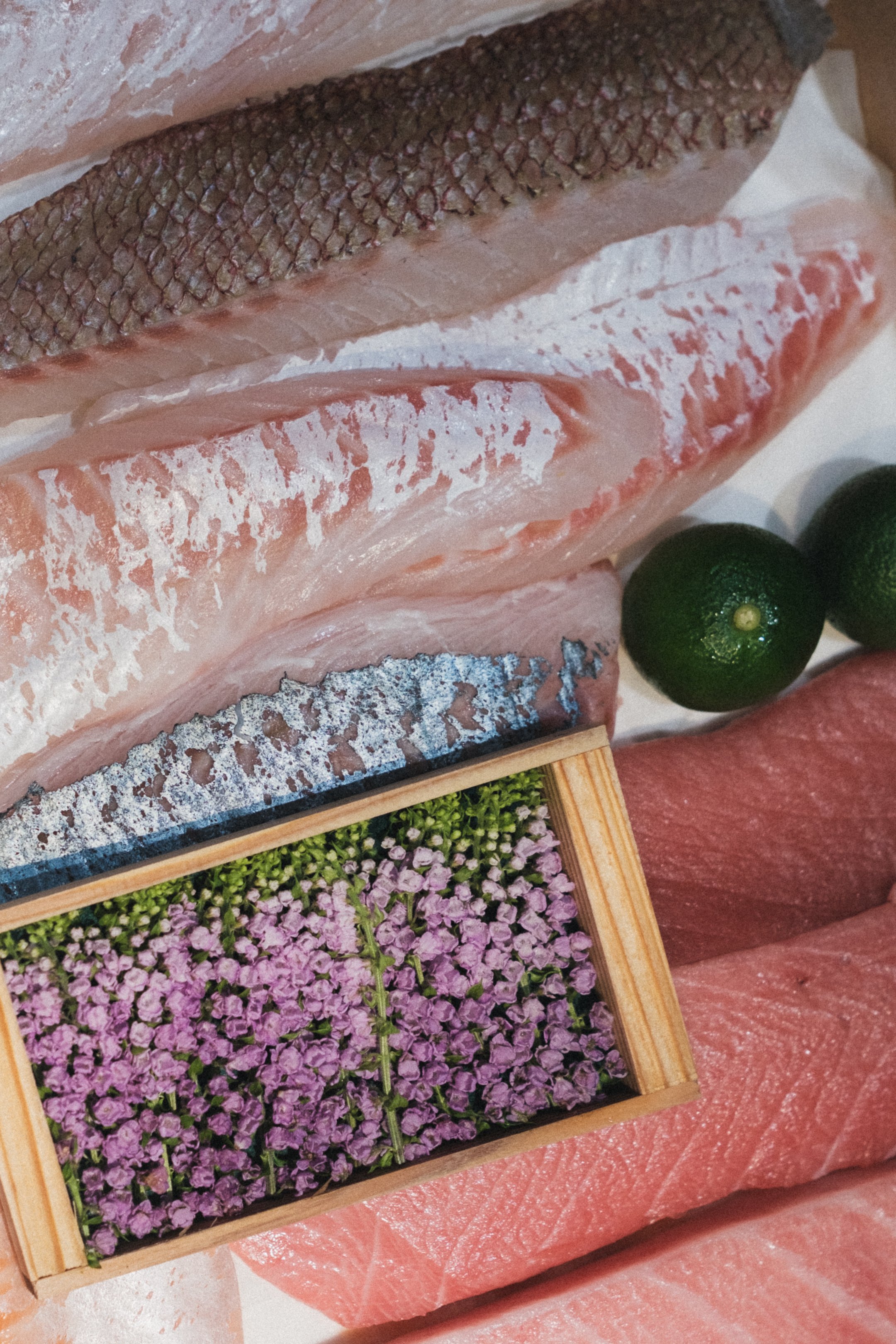 Close-up of raw fish fillets with skin, placed in a wooden box, surrounded by green limes and pink flowers.