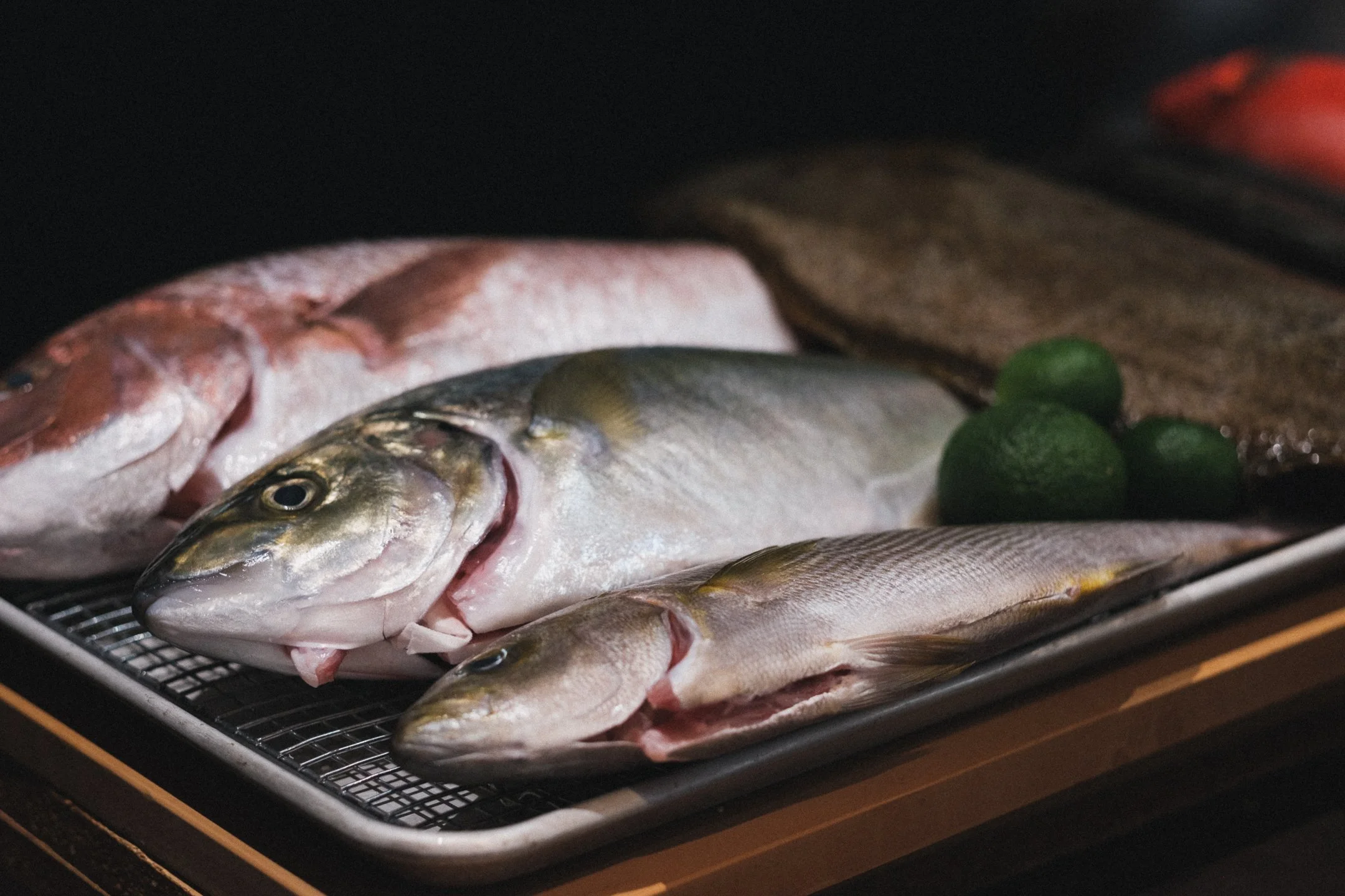 Fresh fish, some whole and some filleted, laid on a tray with green limes in the background.