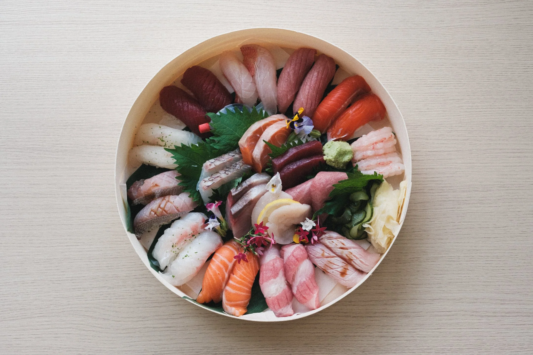 A round platter of assorted sashimi including slices of tuna, salmon, yellowtail, and other fish, garnished with leaves, flowers, and wasabi, placed on a light wooden table.