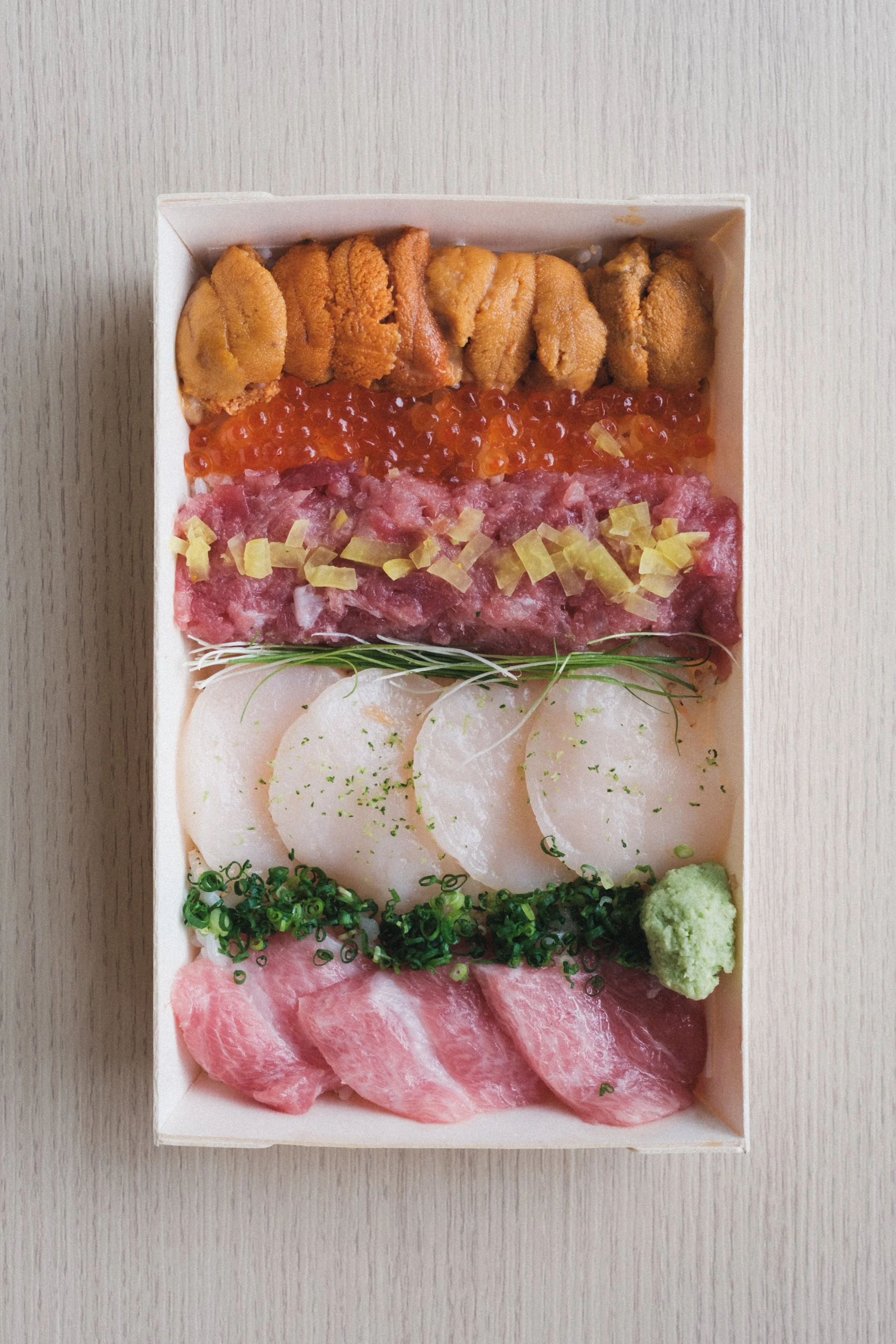 Box of assorted Japanese sashimi and seafood, including various cuts of raw fish, tuna, salmon, and garnishes like wasabi and green onions.