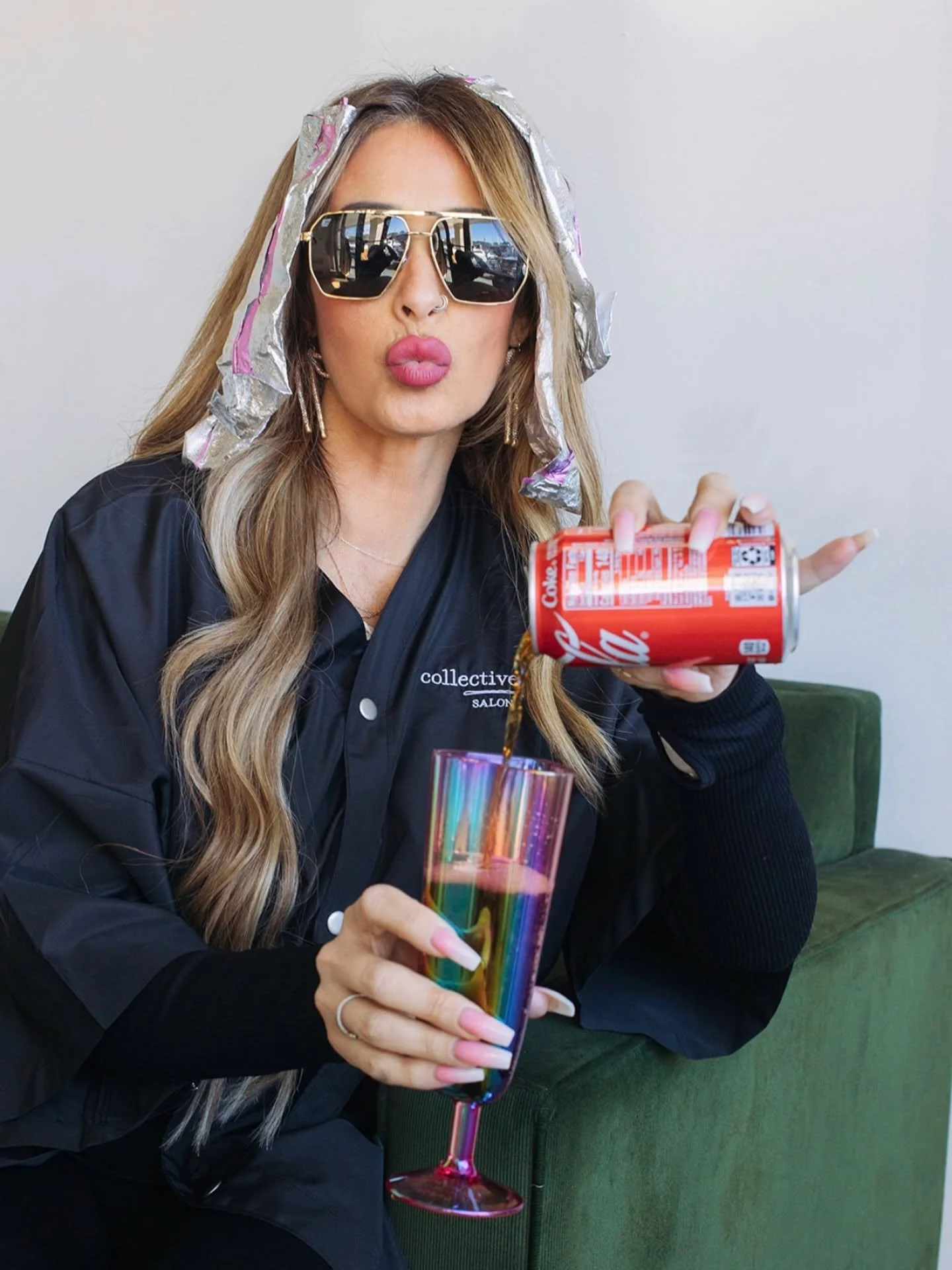 A woman with long wavy hair, wearing large reflective sunglasses and a black jacket, is pouring a dark beverage from a red Coca-Cola can into a tall, iridescent rainbow-colored glass. She is sitting on a green sofa against a plain light-colored wall.