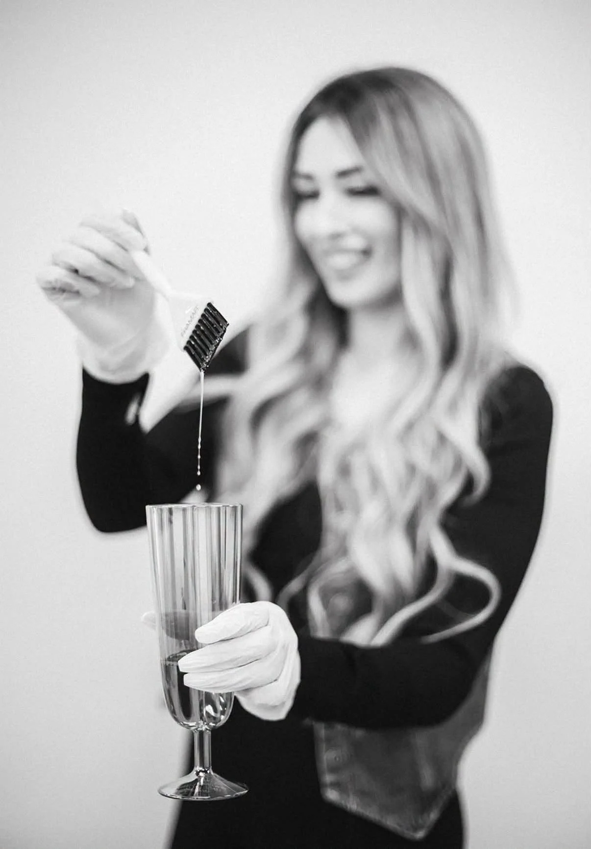 A woman with long, wavy hair wearing a long-sleeved black dress and white gloves, smiling and holding a champagne flute, with a bottle dispenser in her hand over the glass, in black and white.