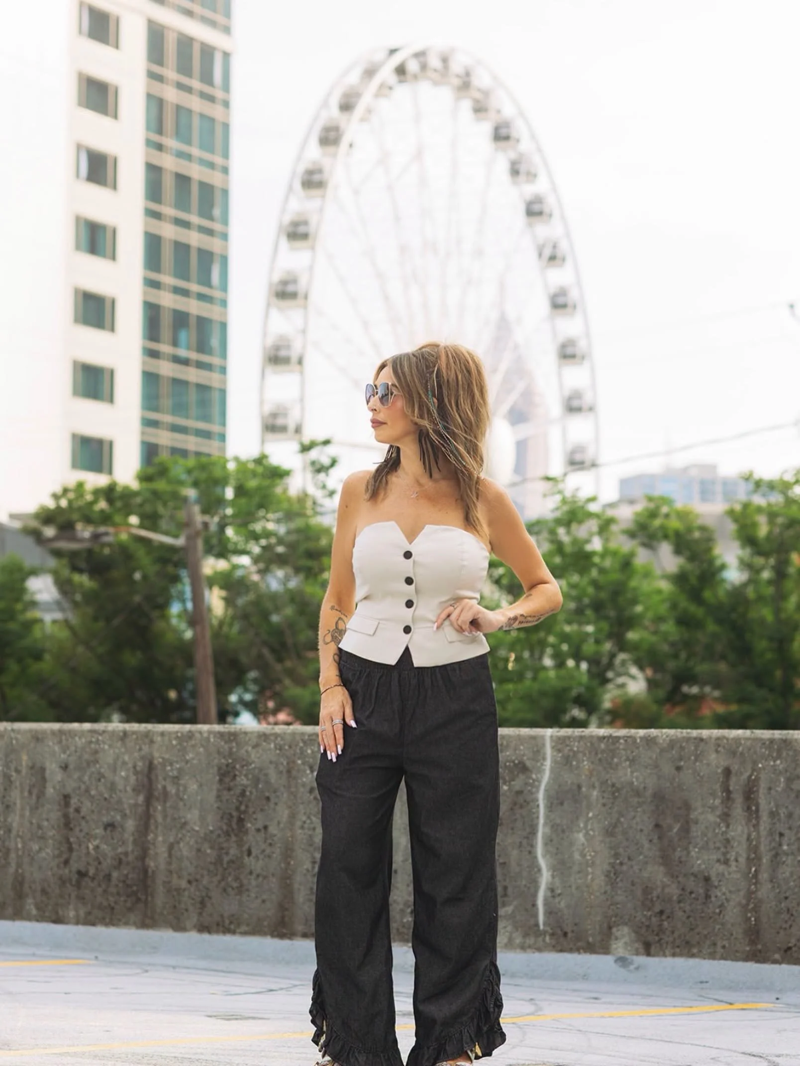 A woman with long hair, wearing sunglasses, a white strapless top with black buttons, and black wide-leg pants, standing outdoors in front of a concrete barrier with a cityscape and a ferris wheel in the background.
