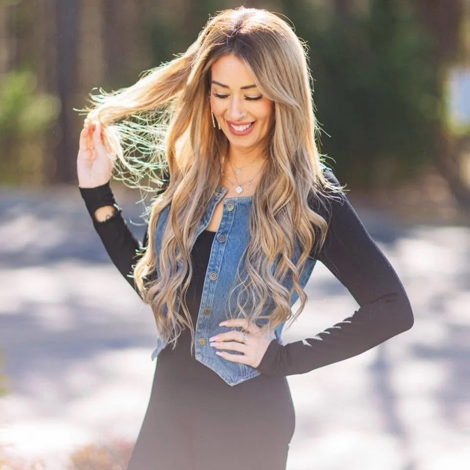 A woman with long, wavy blonde hair smiling outdoors in a natural setting.