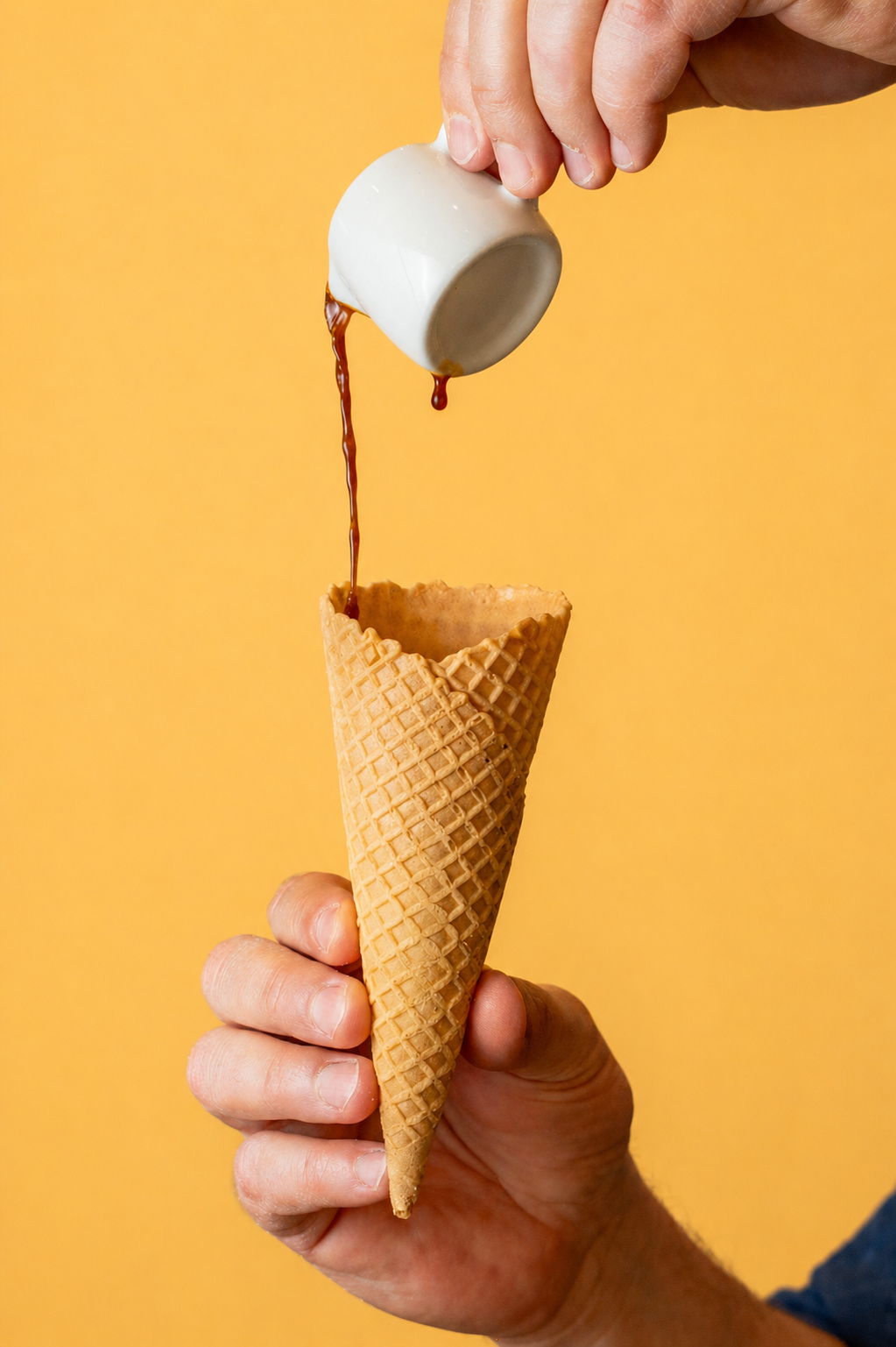 A person holds an ice cream cone with a waffle pattern in front of a yellow background. They are pouring chocolate syrup from a small white cup into the cone.