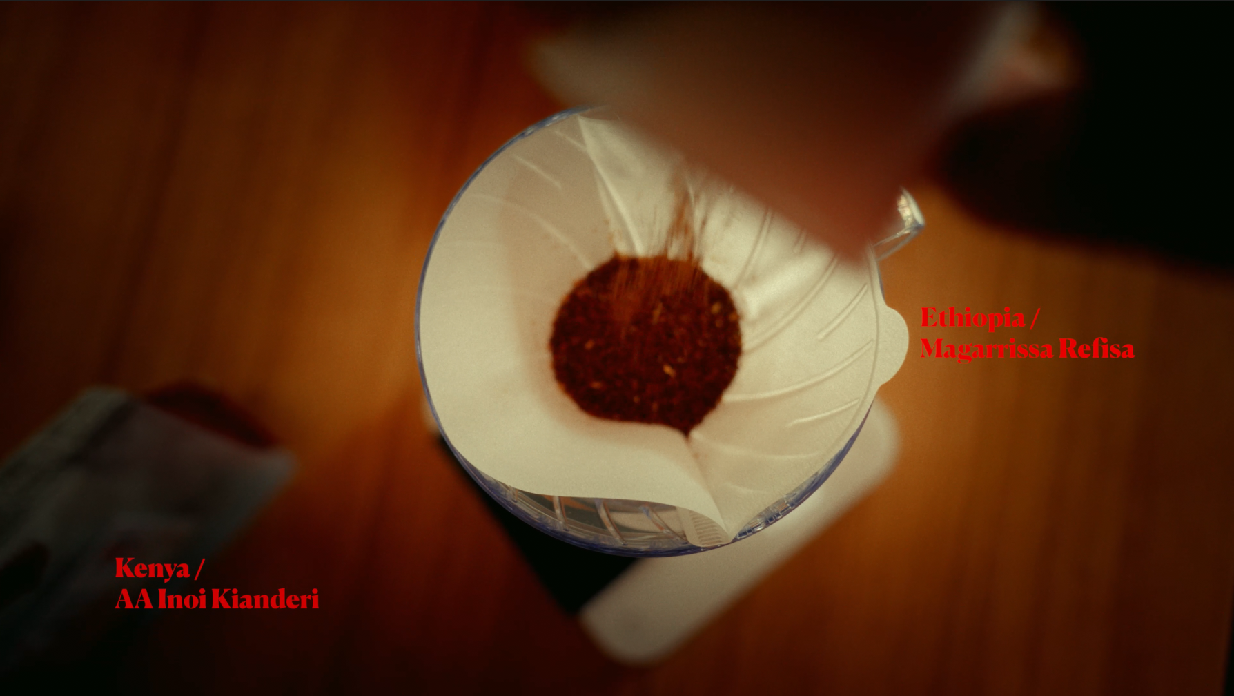Top-down view of a coffee filter with ground coffee inside, placed inside a clear cup. Text overlays indicate Ethiopian coffee from Magarissa Refisa, Kenya AA Inoi Kianderi.