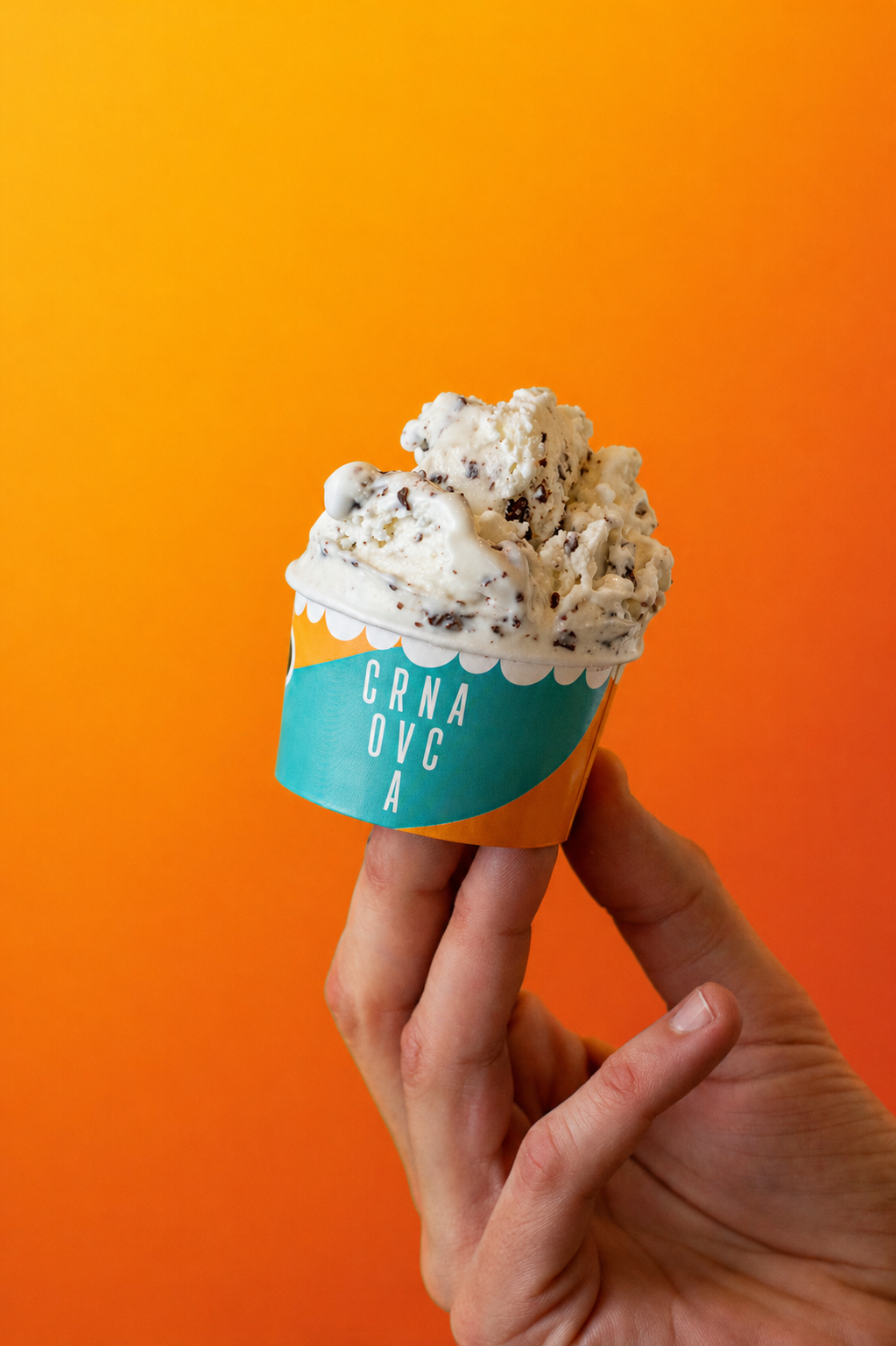 A hand holding a cup of cookies and cream ice cream with chocolate chunks against an orange background.