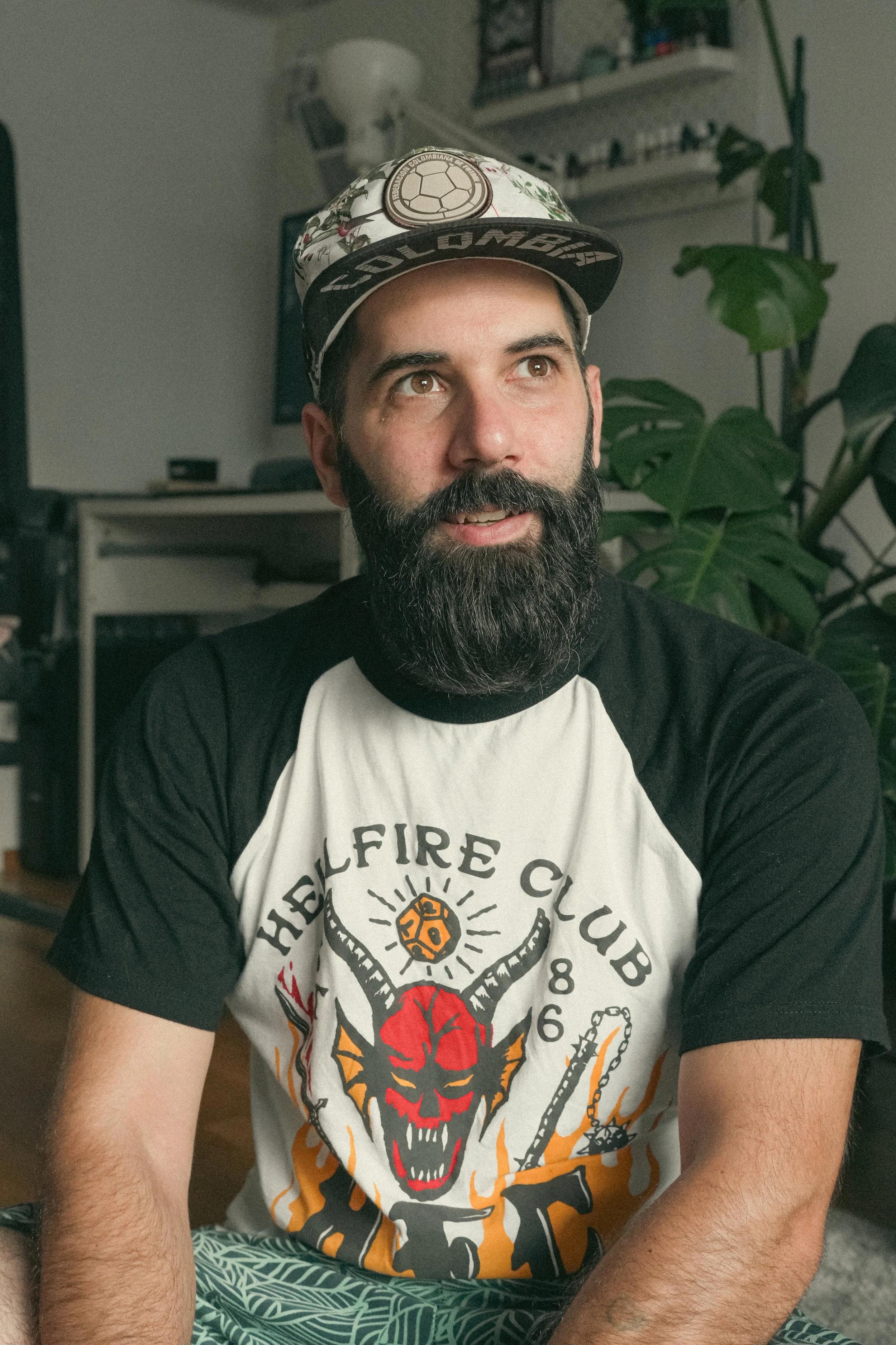 A man with a beard and mustache wearing a floral cap and a black and white T-shirt with a graphic design of a red creature, a yellow sun, and a baseball bat, sitting indoors with plants and shelves in the background.